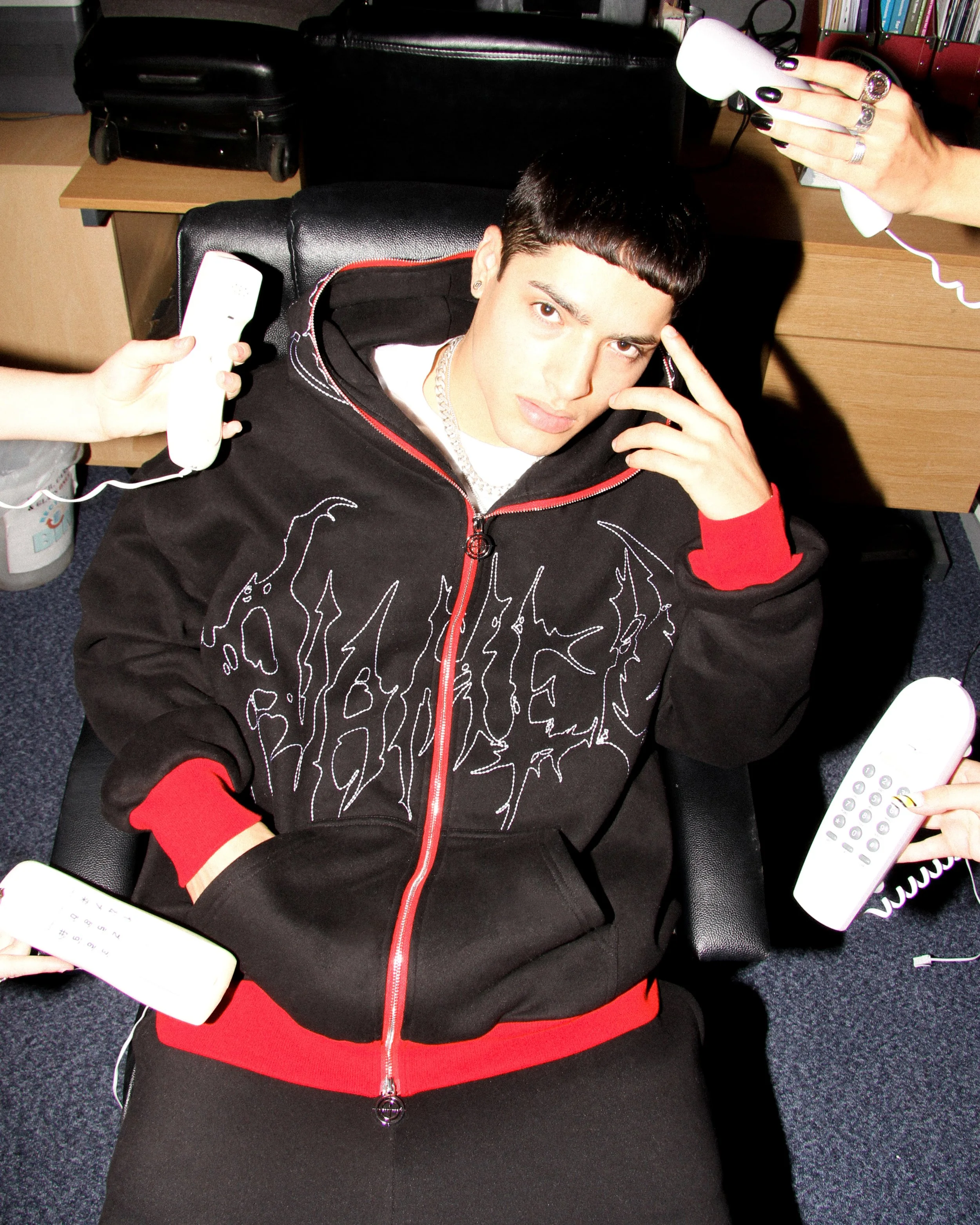 Launch campaign image for NAMED Collective “Classified” showing a model sat in an office chair wearing a black and red hoodie and joggers, surrounded by retro telephones in an editorial setup.