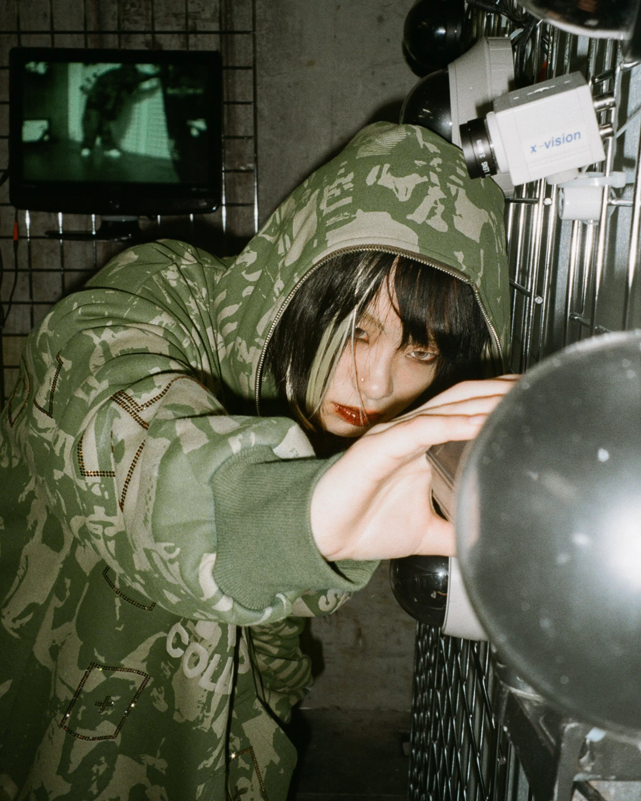 Model in oversized camouflage hoodie reaching toward a surveillance camera in an industrial setting, with CCTV screens and metallic fixtures creating a dark cyber-inspired atmosphere for Named Collective’s “Surveillance” campaign, shot on film.