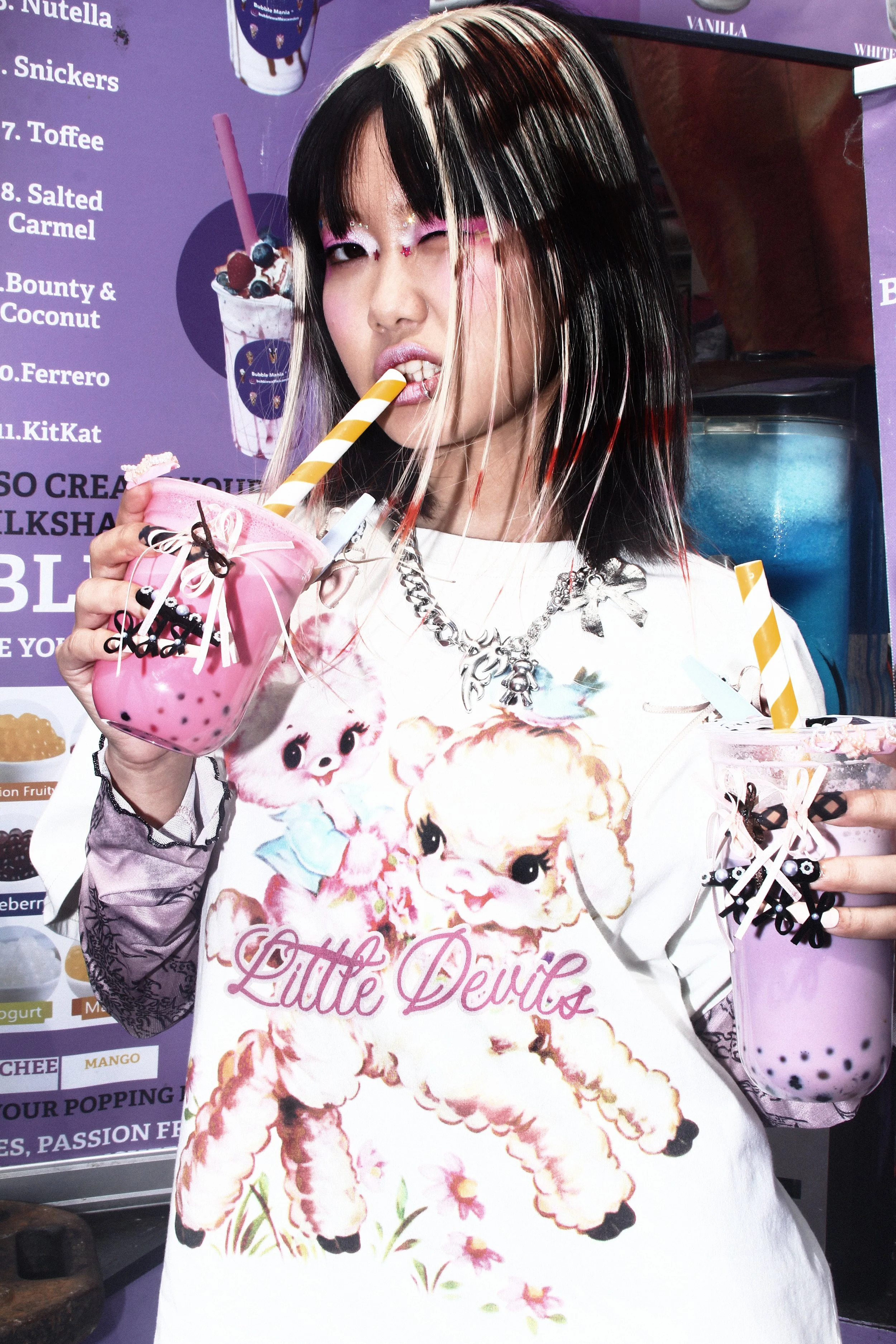 Campaign portrait of a model holding bubble teas while wearing a graphic oversized tee, layered accessories, bold pink eye makeup and star-adorned hair extensions, inside a colourful dessert shop.