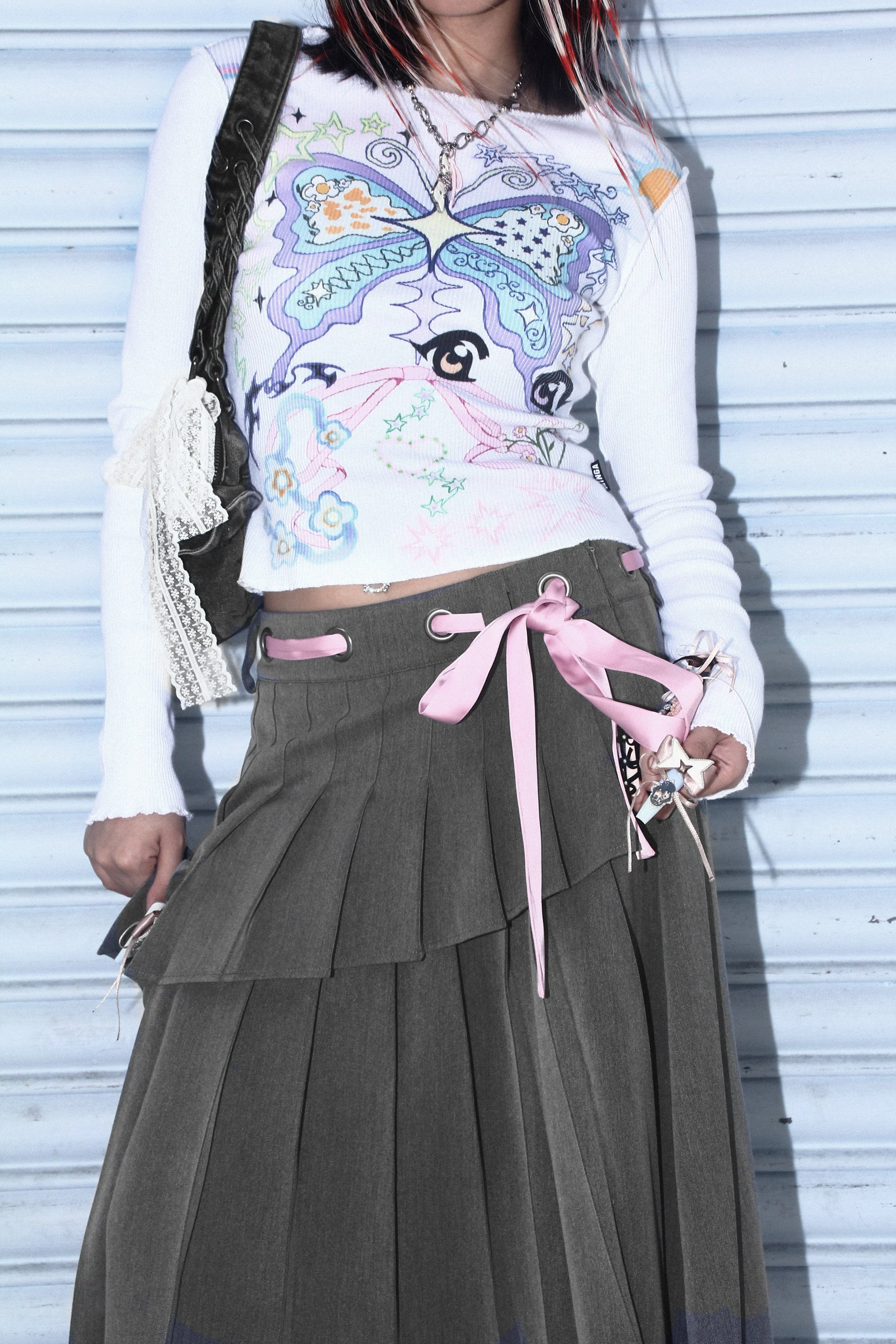 Close-up campaign image featuring a model in a pastel butterfly graphic knit top and layered pleated skirt with pink ribbon detailing, styled with lace accessories and grunge-inspired jewellery against a metallic shutter backdrop.