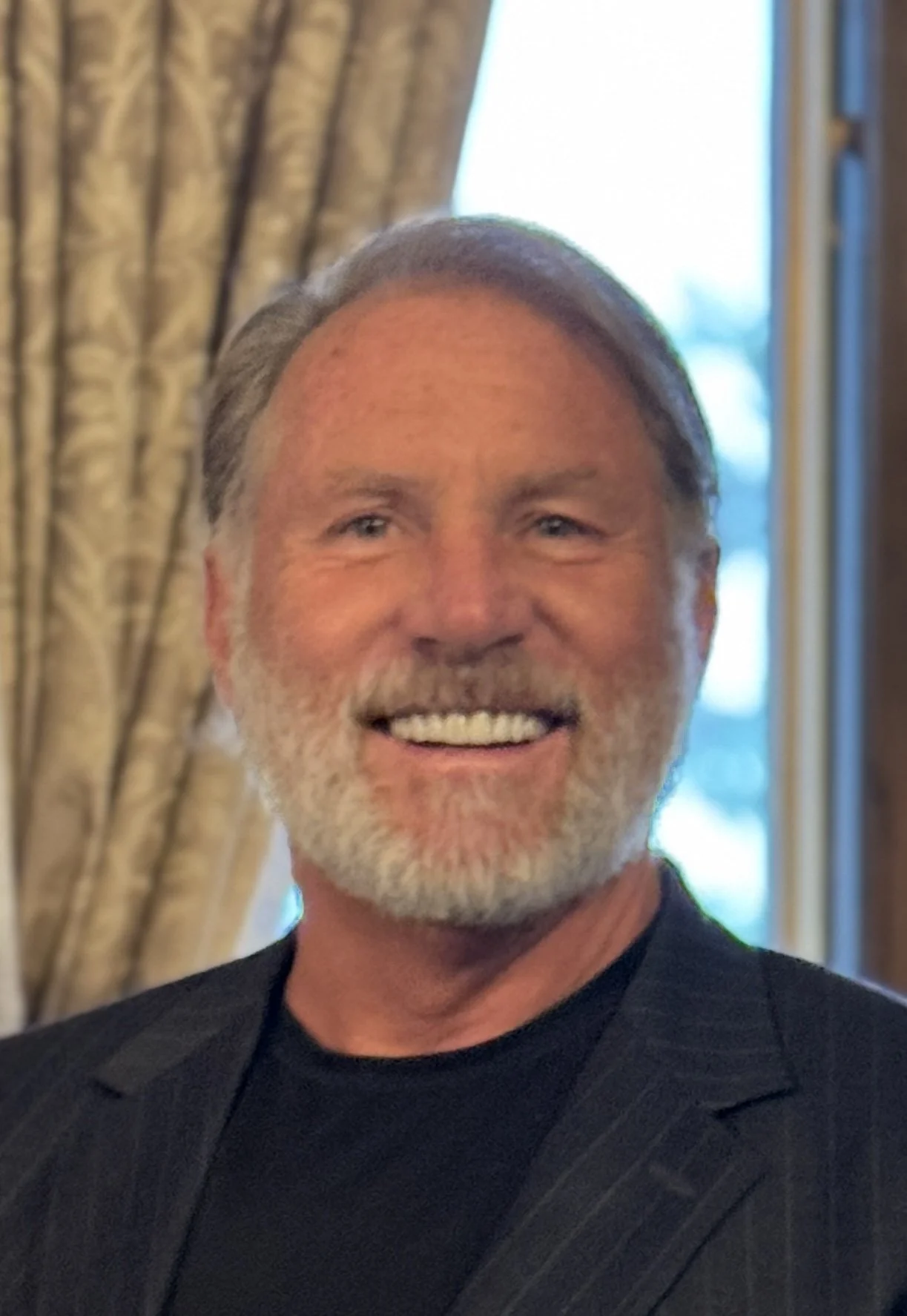 A smiling middle-aged man with gray hair and a beard, wearing a dark blazer and a black t-shirt, standing indoors with curtains and windows in the background.