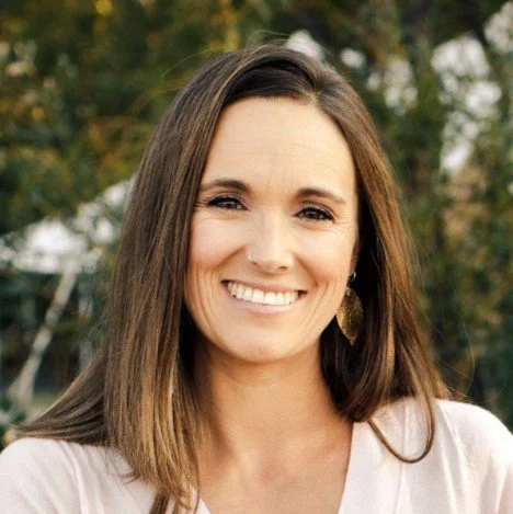 A smiling woman with long brown hair and a nose piercing outdoors, with trees in the background.