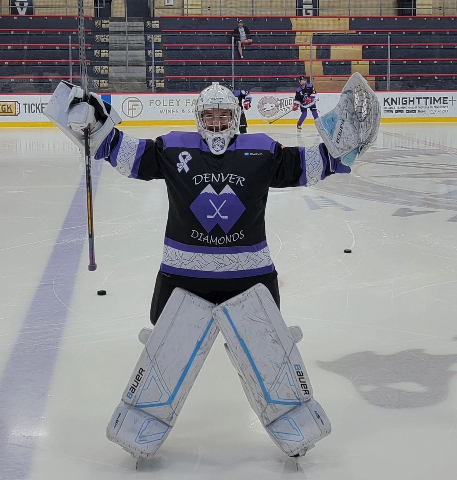 Hockey goalie in black and purple uniform standing on ice rink, holding goalie stick and smiling, with arms raised in victory, in front of other players practicing.