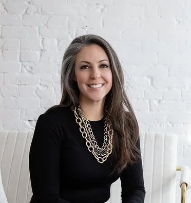 A smiling woman with long brown hair, wearing a black top and layered gold necklaces, sitting in front of a white brick wall.