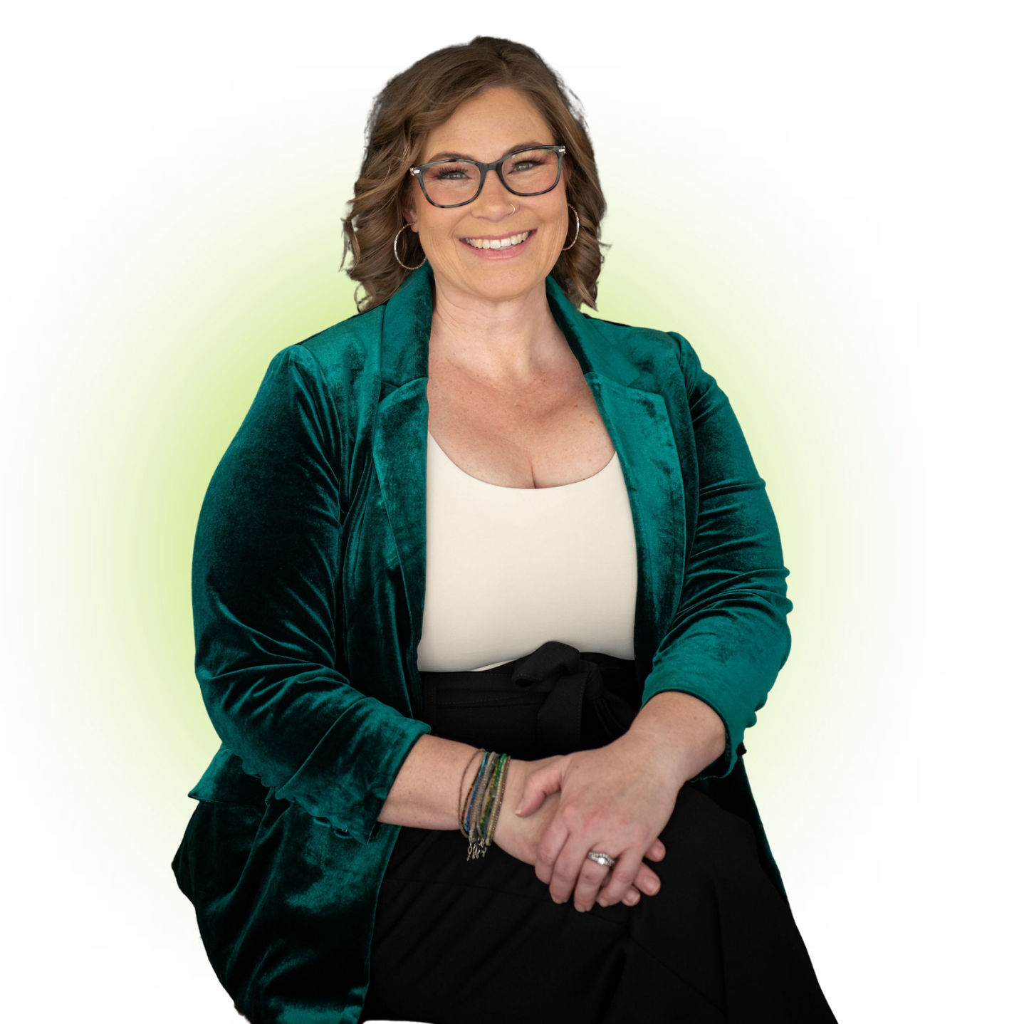 A smiling woman with glasses and wavy brown hair, wearing a teal velvet blazer, white top, and black skirt, sitting with her hands clasped, against a green glowing background.