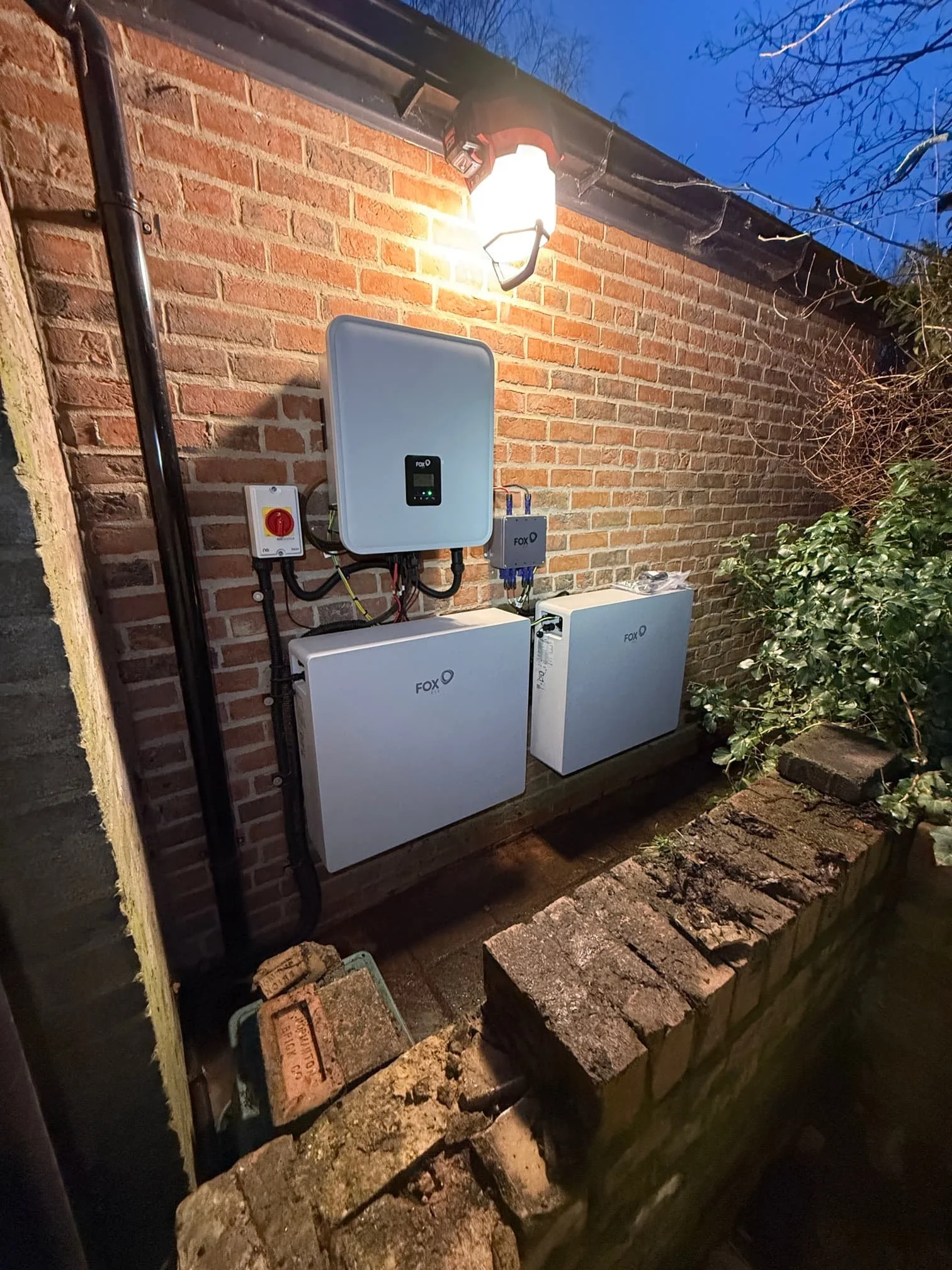 Installation of solar power equipment on a brick wall at dusk, including white control units, wiring, and an outdoor light fixture.