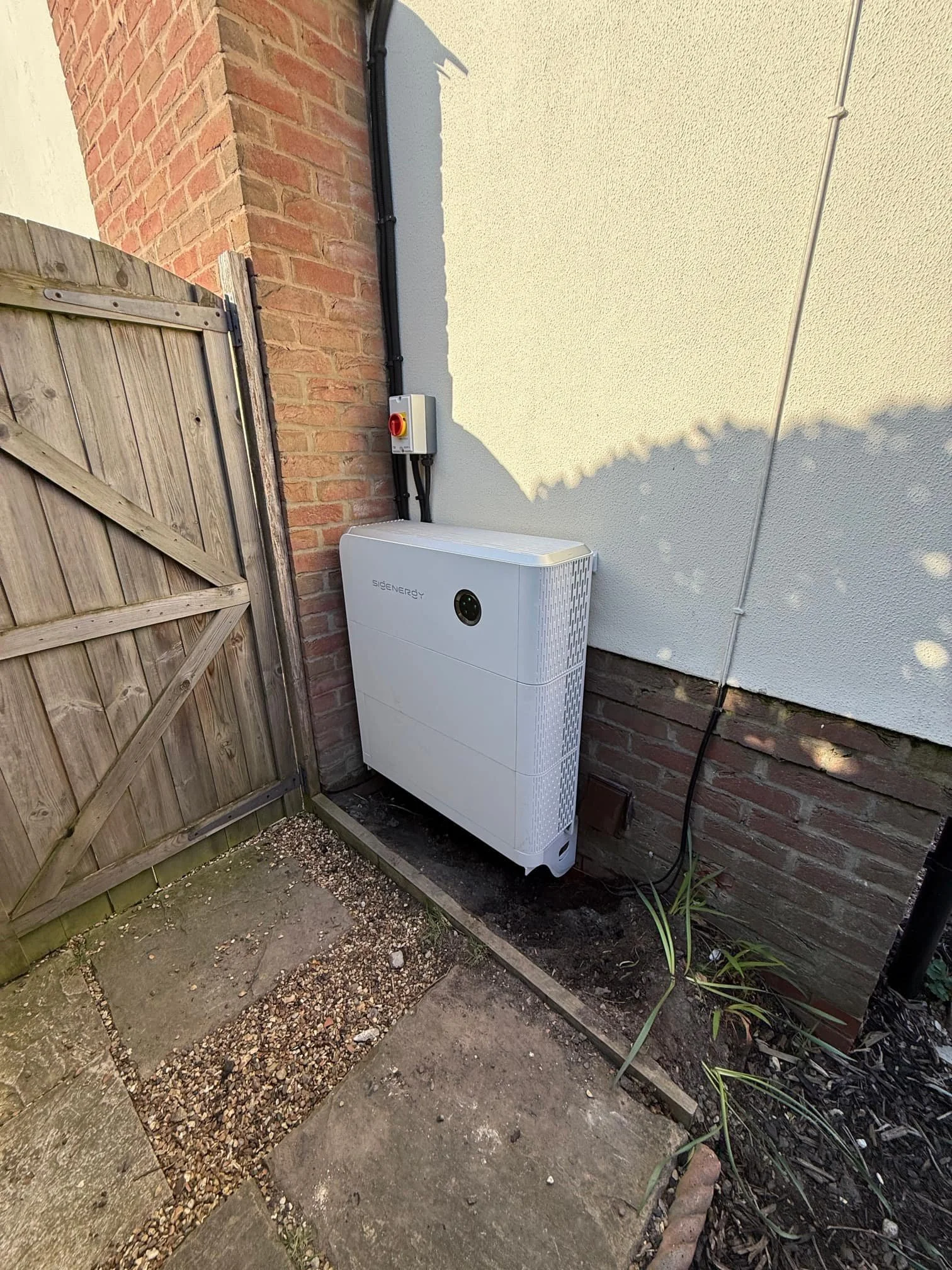 An outdoor area next to a brick wall and a white wall, featuring a wooden fence, a large white modern appliance with the brand name 'SigEnergy', electrical cables attached to a control box, and some plants and gravel on the ground.