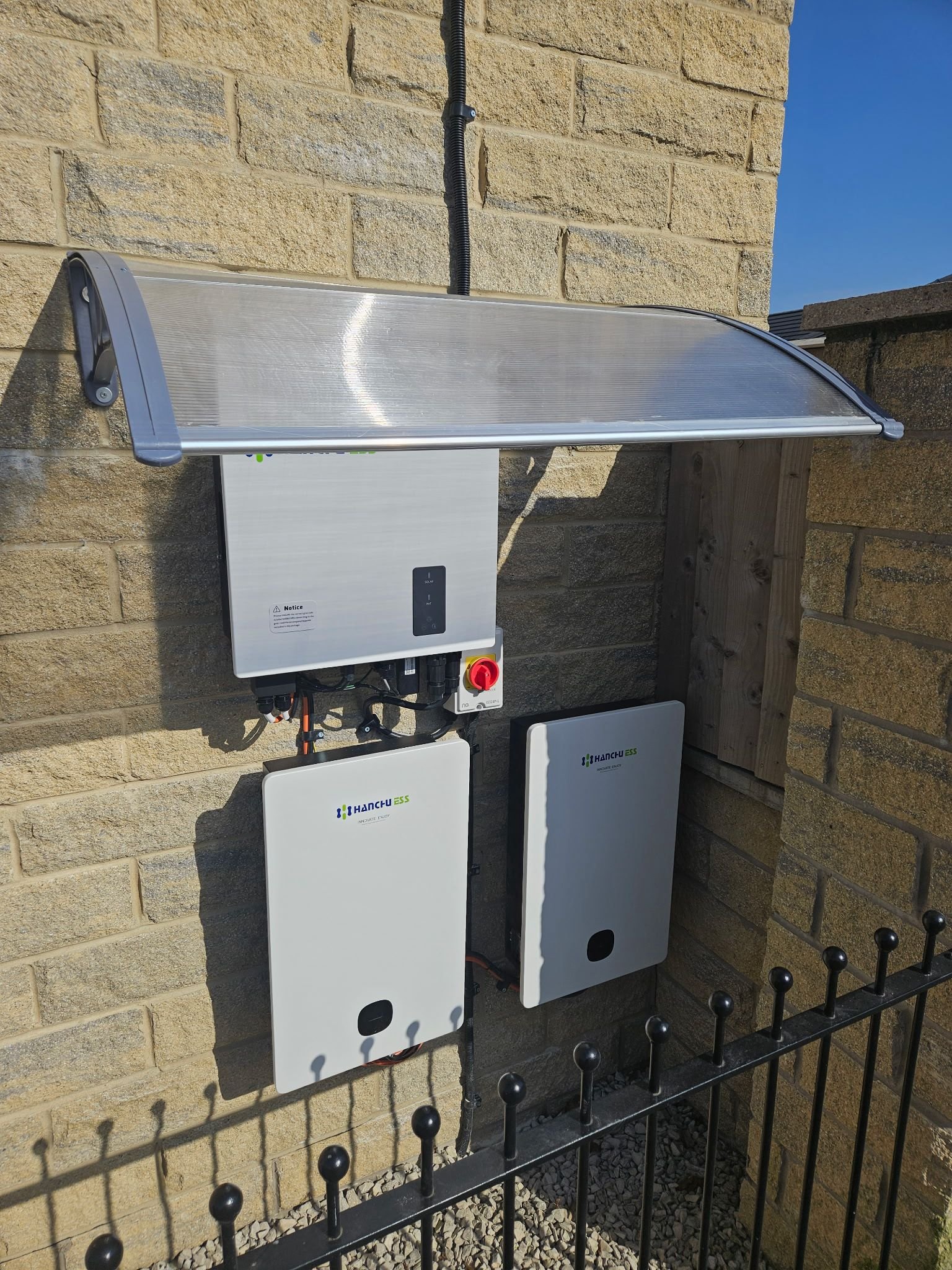 Outdoor solar power system with two white units, a solar panel cover, and electrical wiring installed on a brick wall, with a black metal fence below.