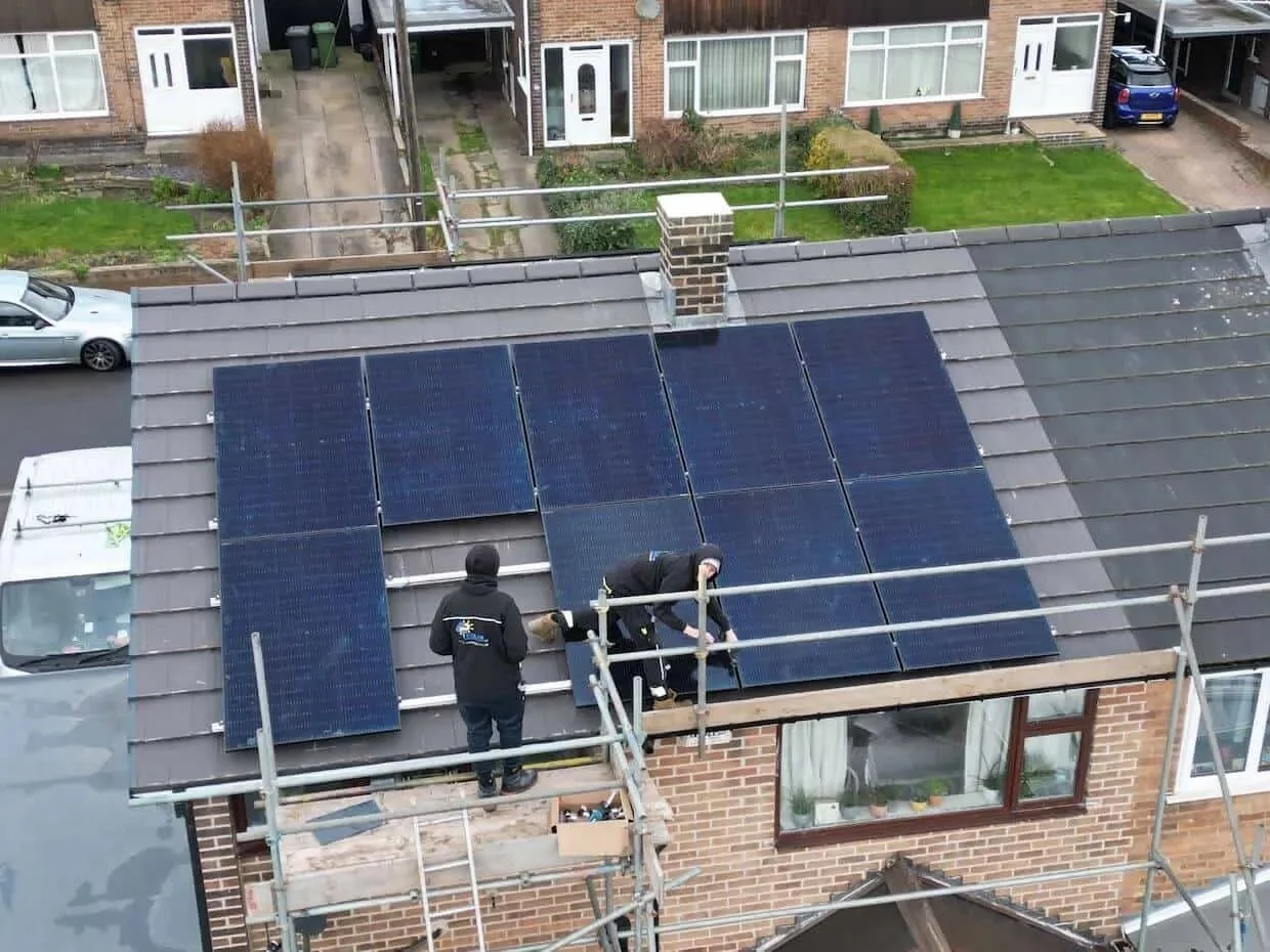 Solar panel installation in progress on a residential roof, with installers fitting a multi-panel solar array around a chimney using scaffolding for safe access.