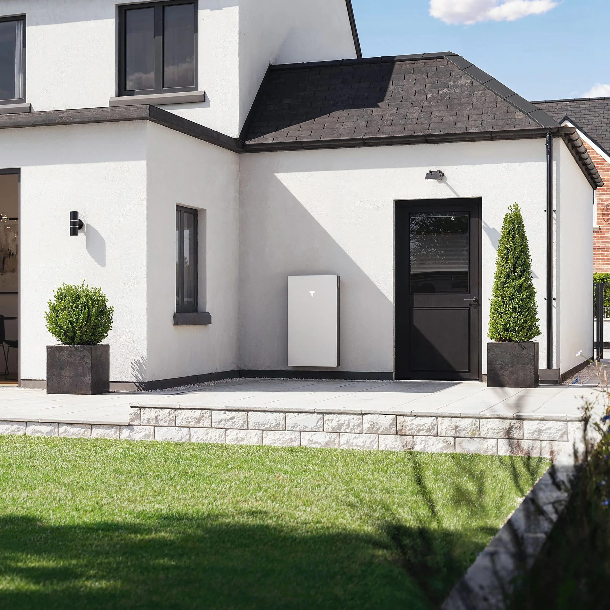 Modern two-story house with white walls, black window frames, black door, and a dark tiled roof, with potted bushes and a grassy lawn in front.