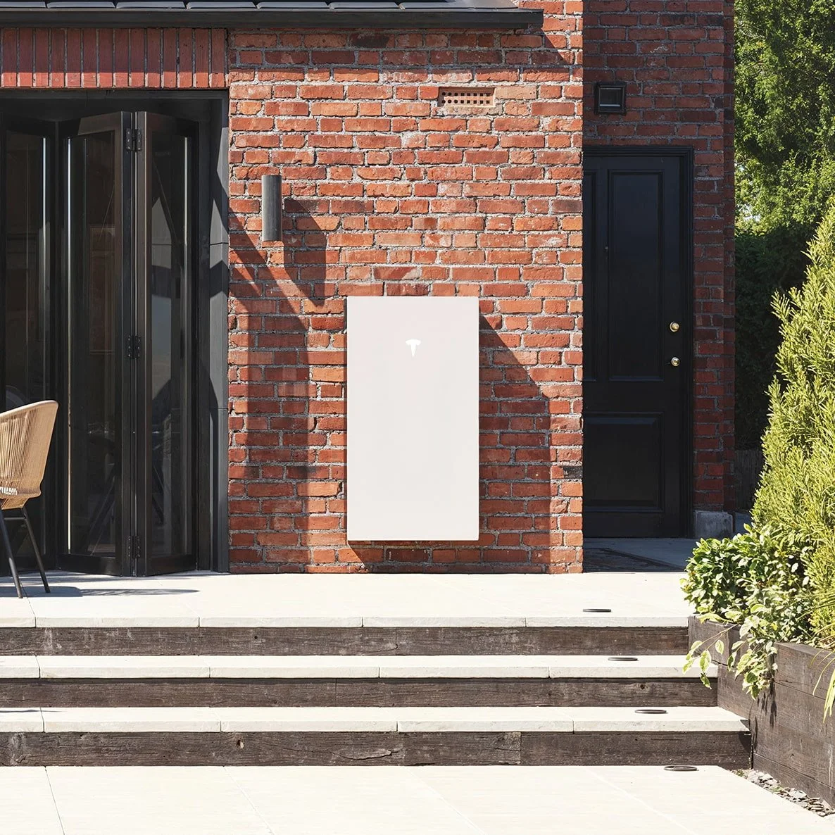 Red brick house exterior with black door, white utility box, small black wall-mounted light, and planters with green bushes on the steps outside.