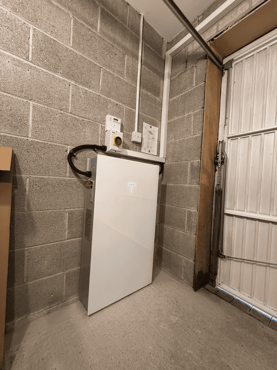 Tesla Powerwall energy storage unit mounted on a concrete wall in a garage, near electrical panels and a garage door.