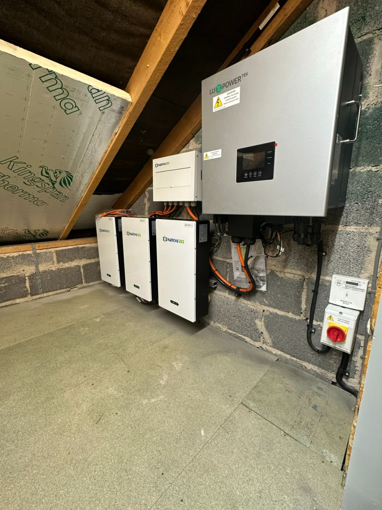 Indoor view of an electrical setup with a gray solar power inverter and two black and white batteries mounted on the wall in a room with brick and wood walls, and a gray carpeted floor.