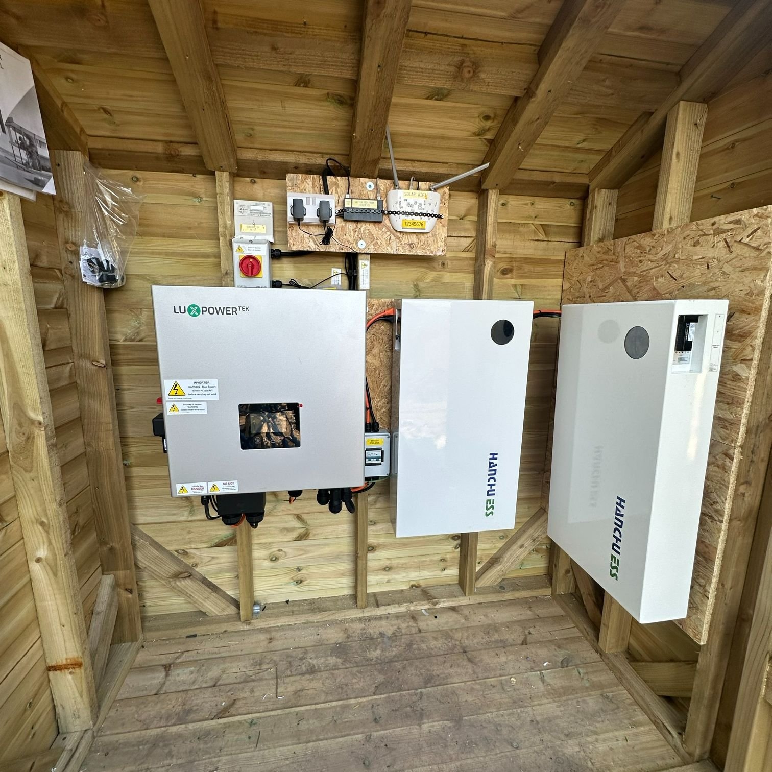 The image shows a wooden structure housing a solar power system, including an inverter, two white battery units, and electrical components mounted on the wall with wiring connected between them.