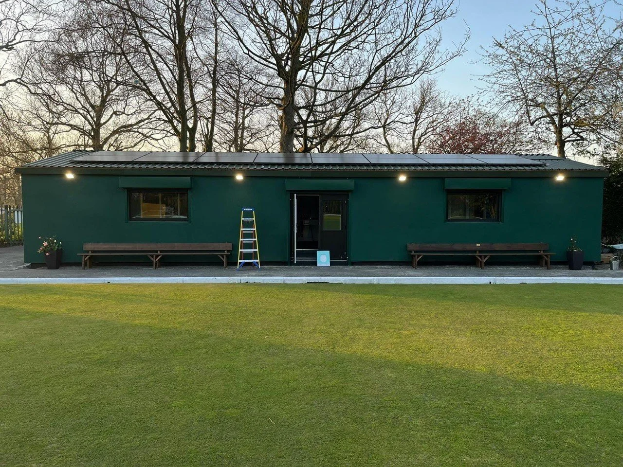 Single-story green building with a metal roof, surrounded by grass and trees. There are two benches outside, a ladder leaning against the building, and potted plants on either side.