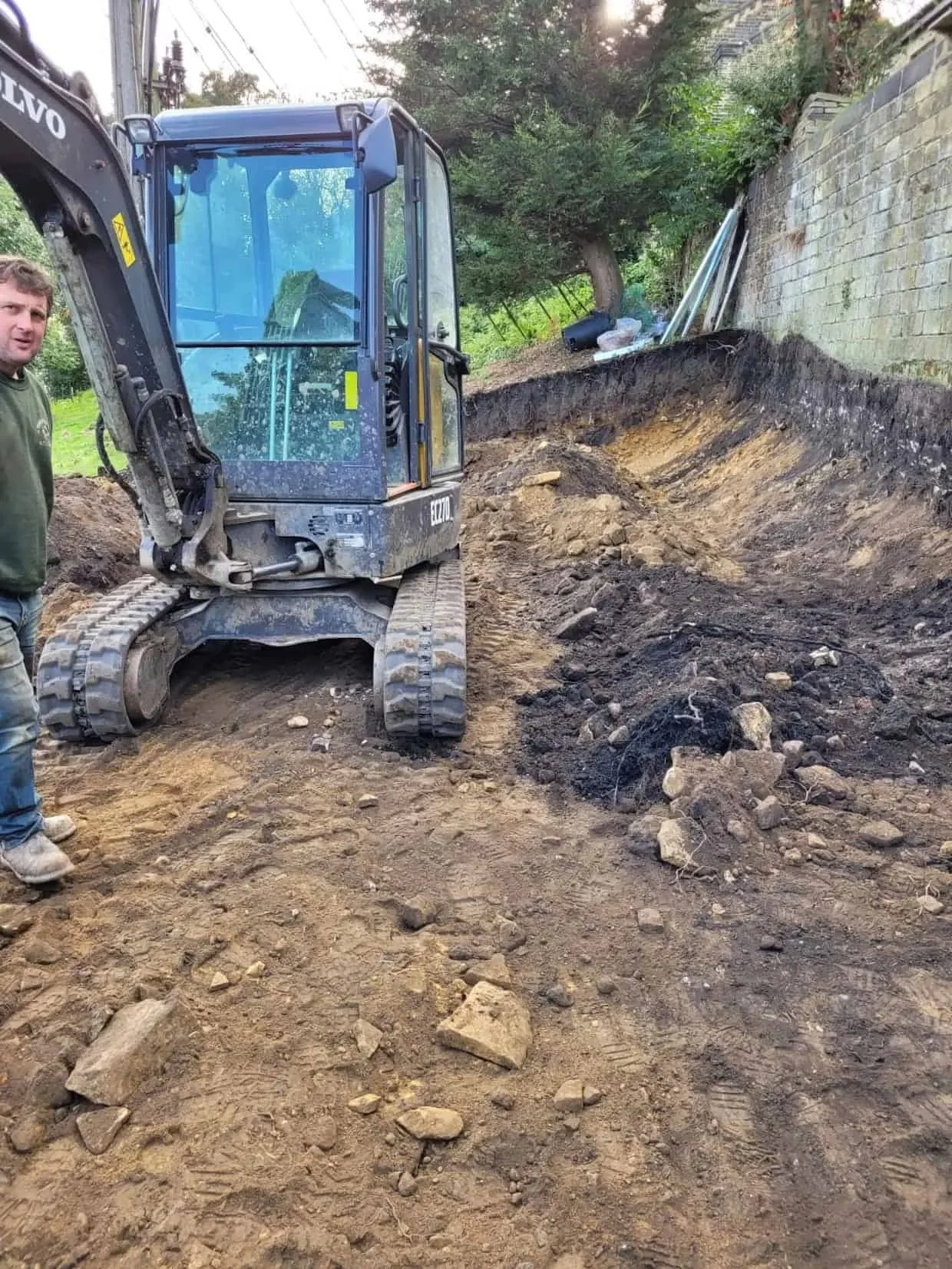 Excavation work underway for a ground-mounted solar panel installation, with a mini digger preparing foundations and cable trenches in a residential garden.