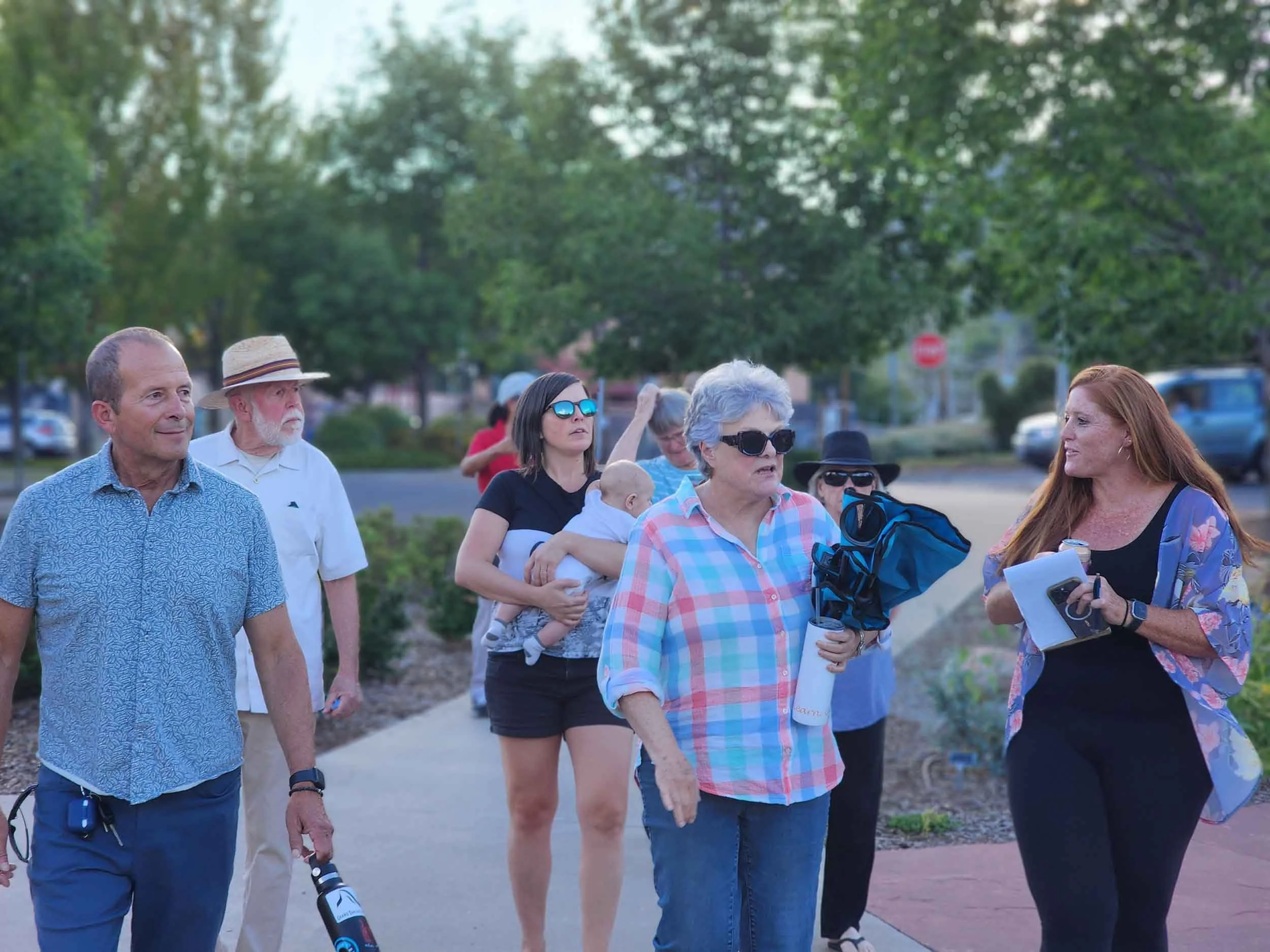 Group of seven people walking outdoors on a sidewalk, engaged in conversation. There are women and men of various ages, some wearing sunglasses, with trees and parked cars in the background.
