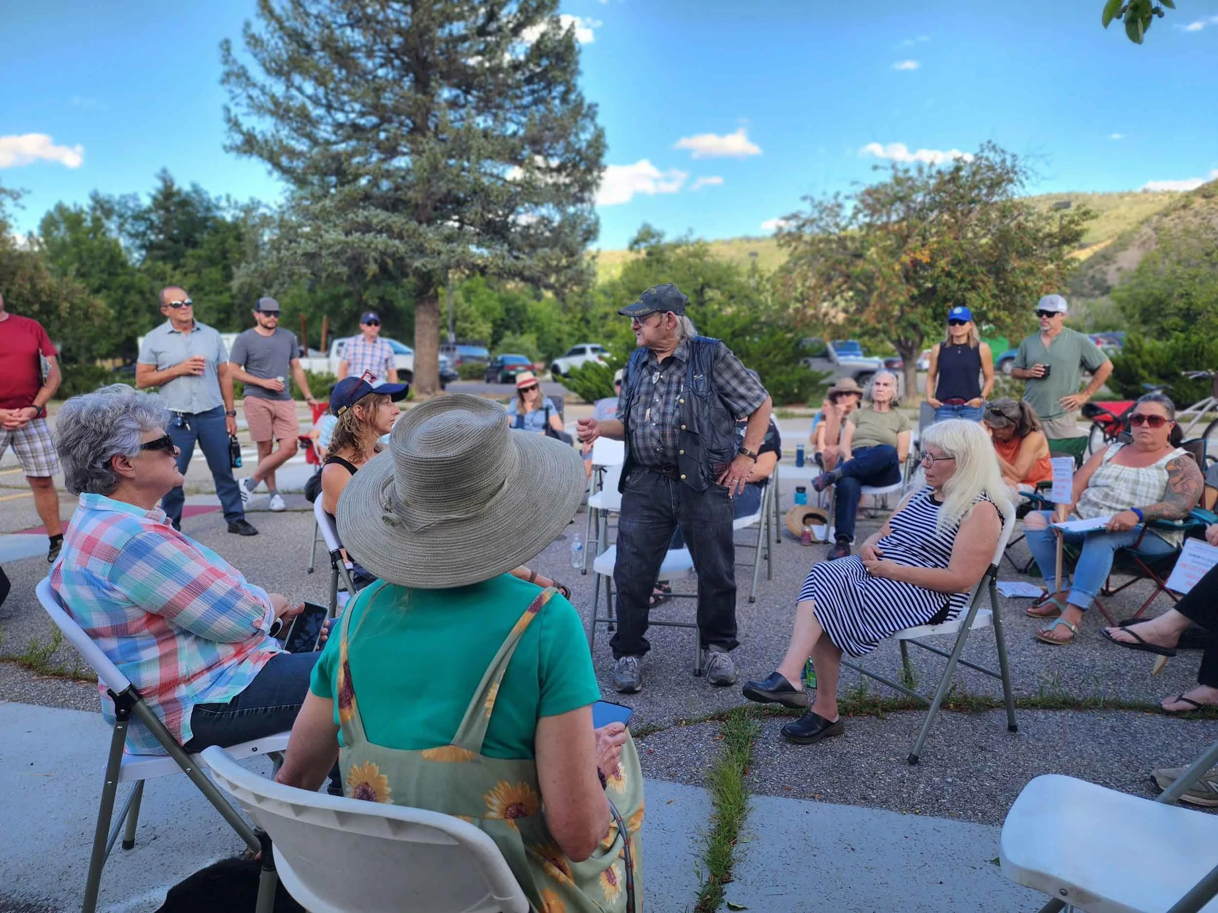 Group of people gathered outdoors during daytime, some seated and some standing, with a man speaking to the group in front of trees and parked cars.