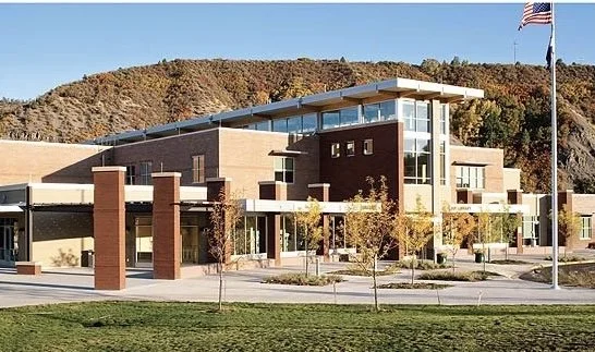 Modern school building with glass windows, brick walls, and a landscaped courtyard with trees, flagpole, and American flag, set against a hillside with autumn foliage.
