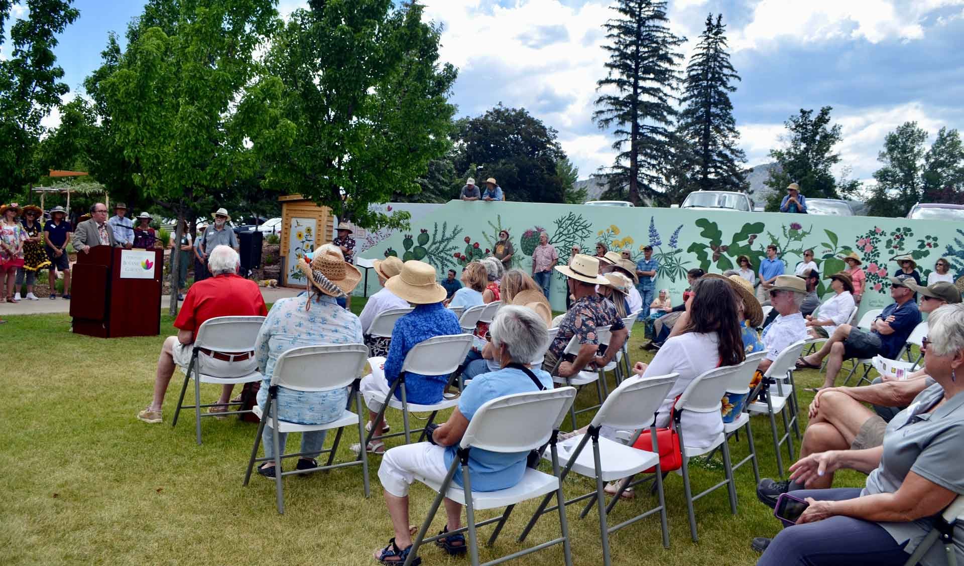 Outdoor gathering with people seated on white chairs, attending a presentation or speech, with some standing at a podium and others on a stage, in a garden setting with trees and a decorative wall with plant illustrations, under partly cloudy skies.
