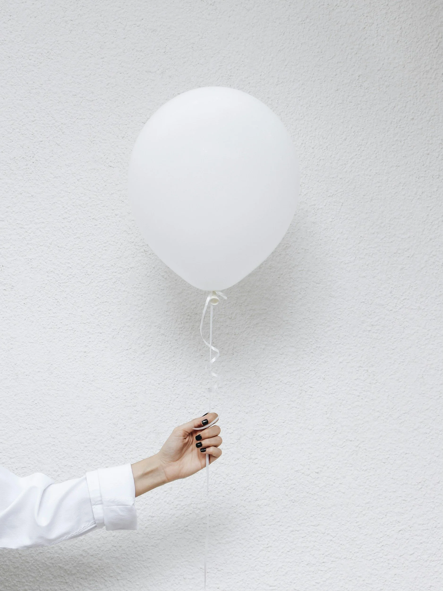 A person wearing a white sleeve holds a white balloon against a light textured wall.