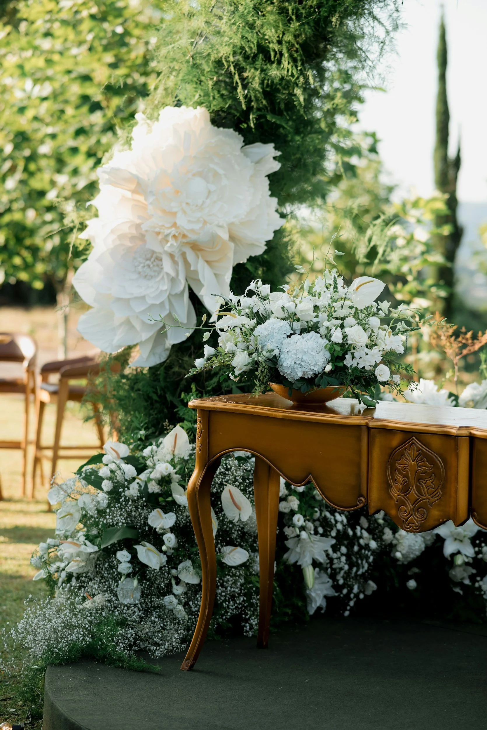 White floral arrangement on a wooden table with green foliage and chairs in the background, outdoors during daytime.