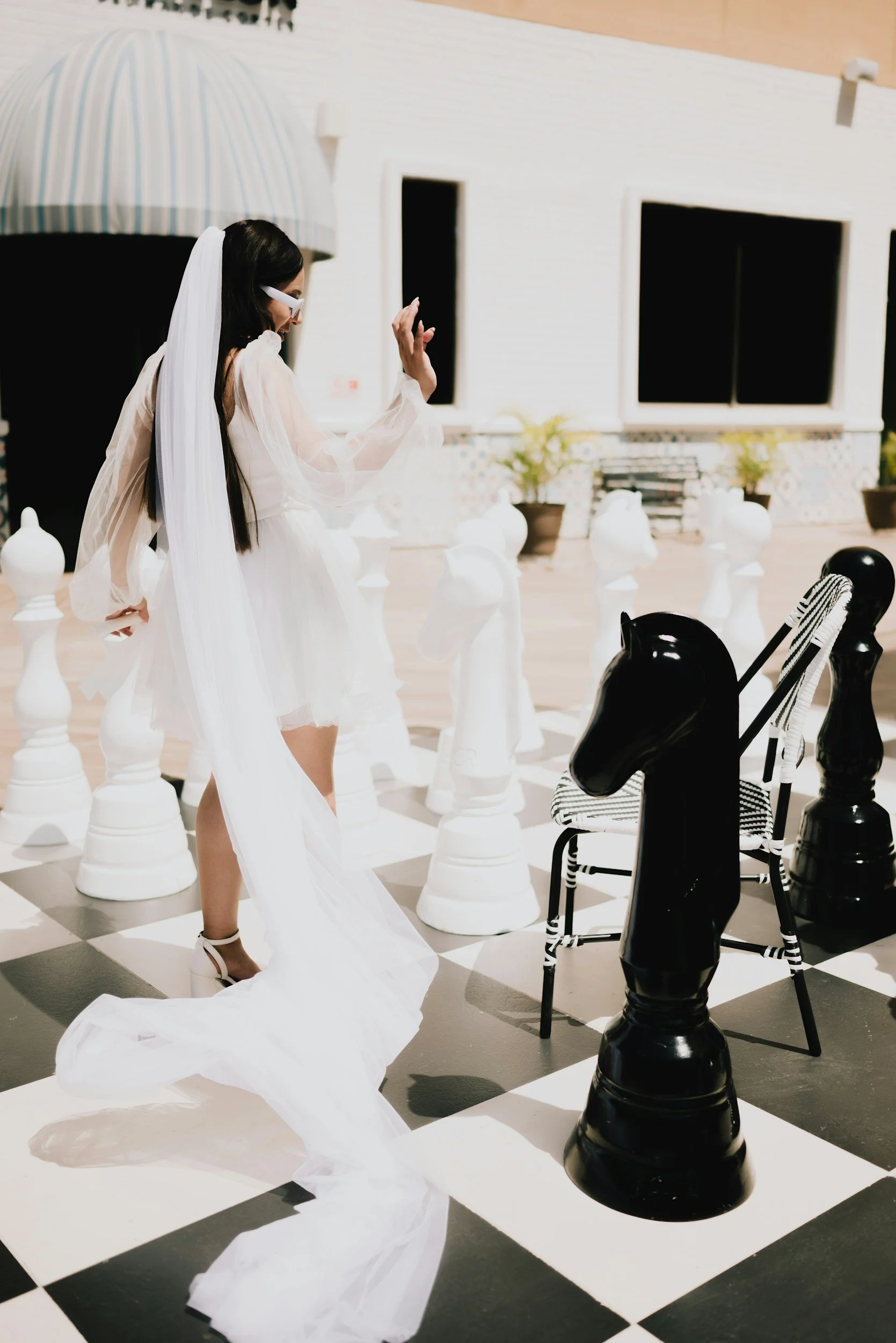A woman in a white dress and veil playing chess on an outdoor black-and-white checkered chessboard.