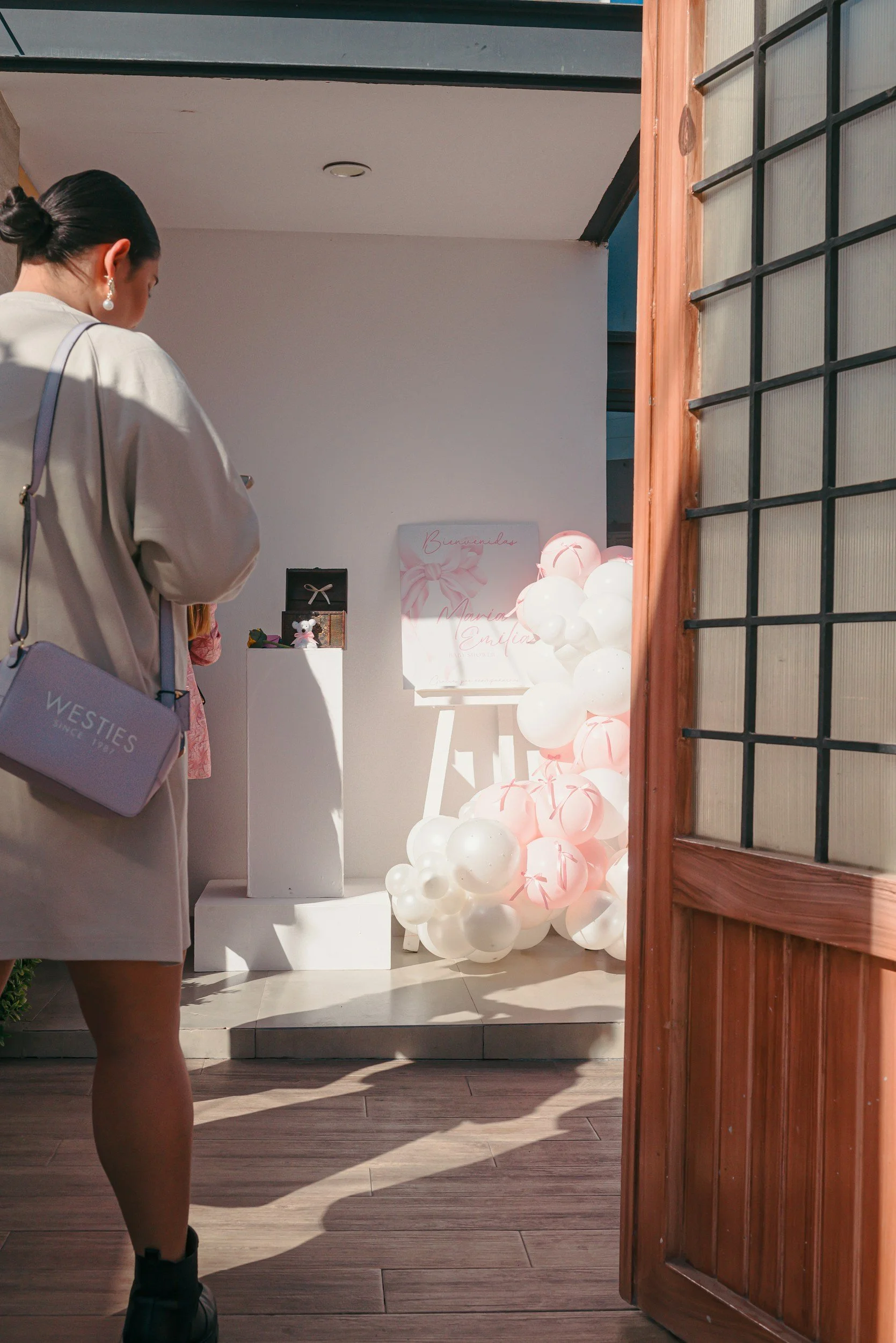 A woman in a beige jacket and black shoes standing inside a room, looking at a display with white and pink balloons and a sign that reads 'Bienvenida Mariana Emilia'.