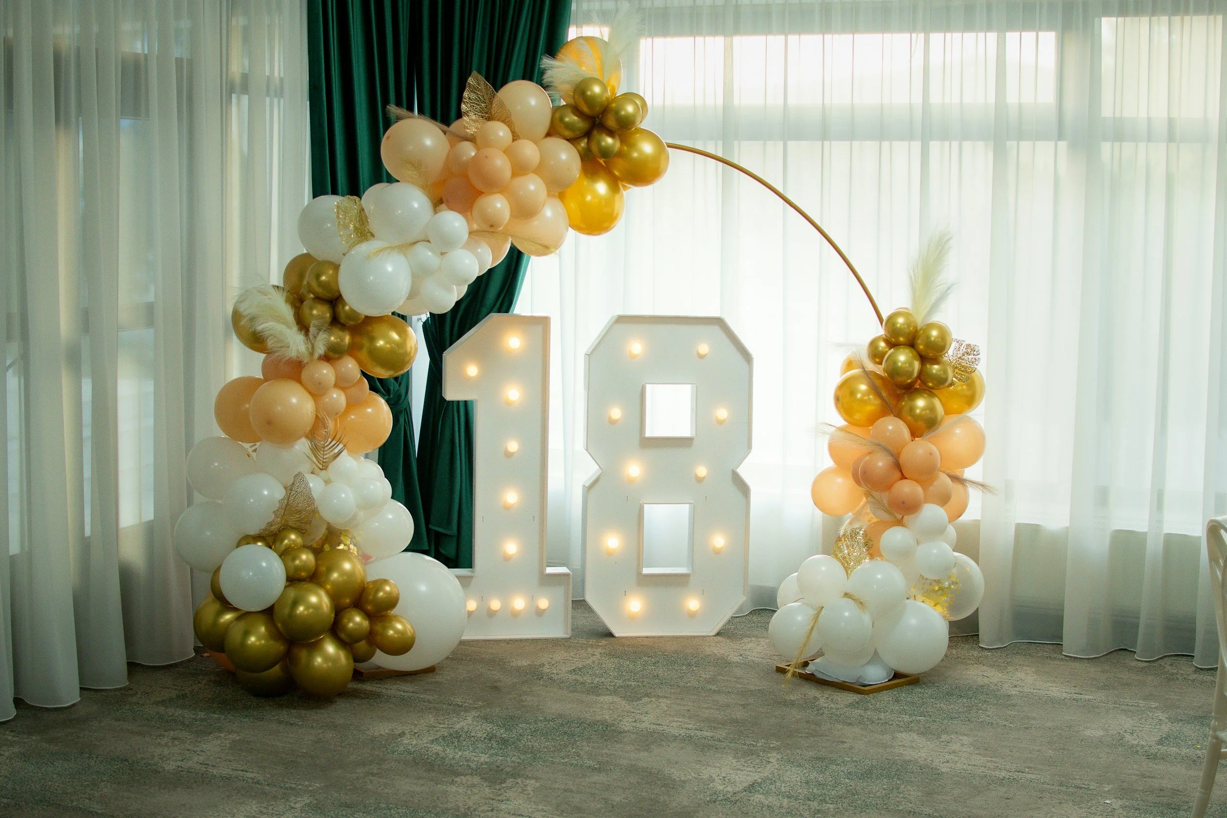 Decorative display with large illuminated number 18 surrounded by balloon bouquets in gold, white, and peach colors, with some feathers and glitter accents, set against sheer curtains and a green backdrop.