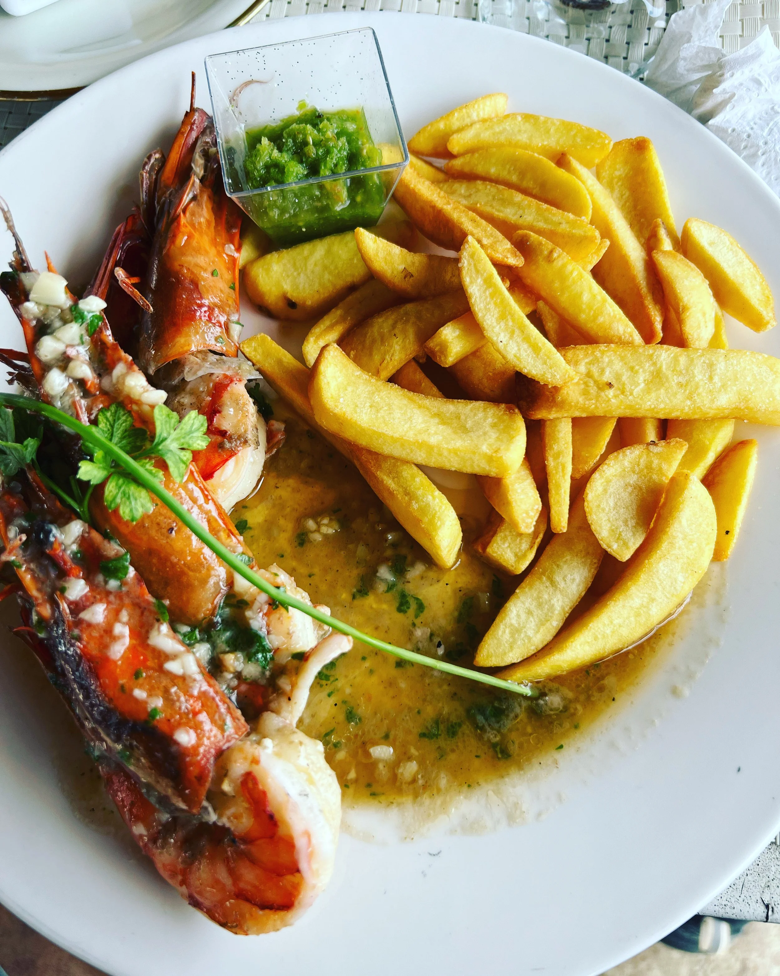 Lobster tail with herbs, served with French fries, green sauce, and melted butter on a white plate.