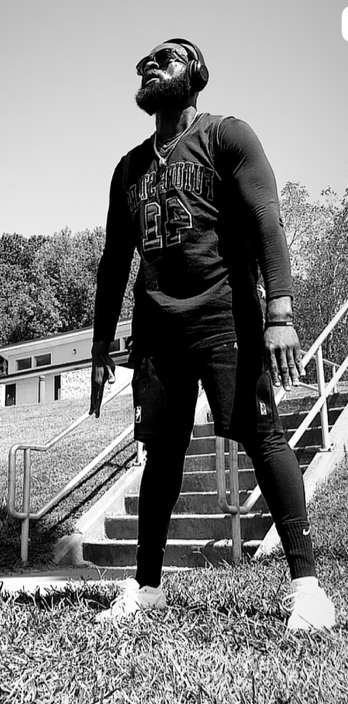 A man standing outdoors on grass, wearing headphones, sunglasses, and sportswear, with a building and trees in the background.