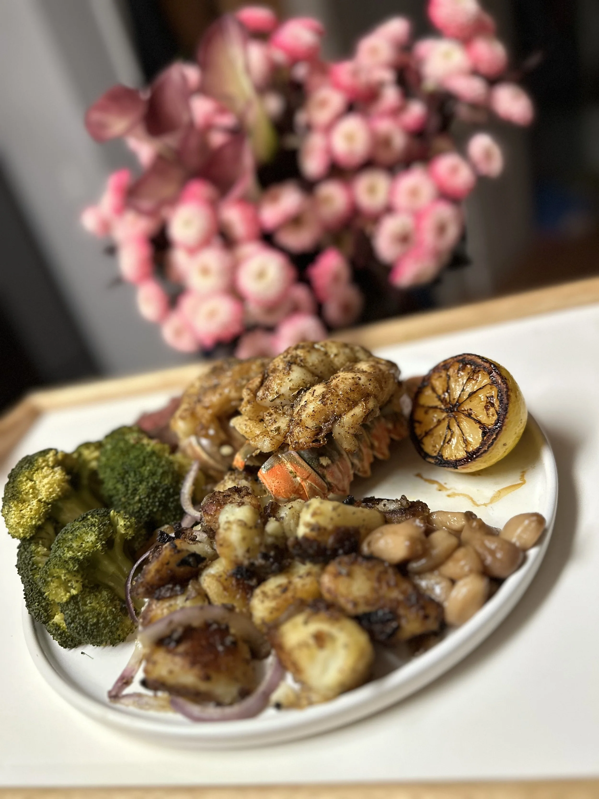 Plate with grilled broccoli, shrimp, grilled lemon, vegetables, and beans, with blurred pink flowers in the background.