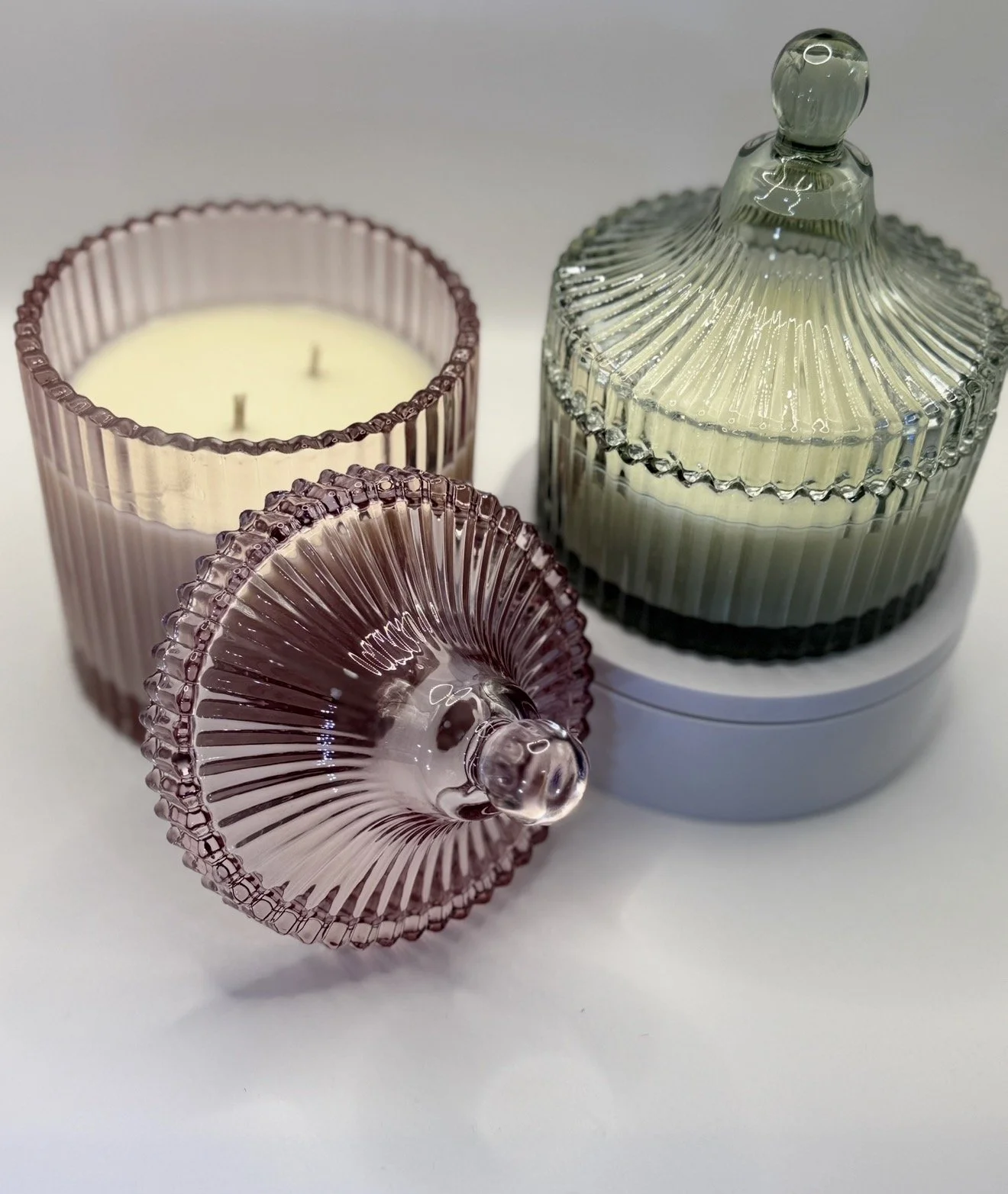 Decorative glassware set including a pink ribbed candle holder with a white candle, a pink ribbed perfume bottle with a round stopper, and a large ribbed glass jar with a lid, all on a white surface.