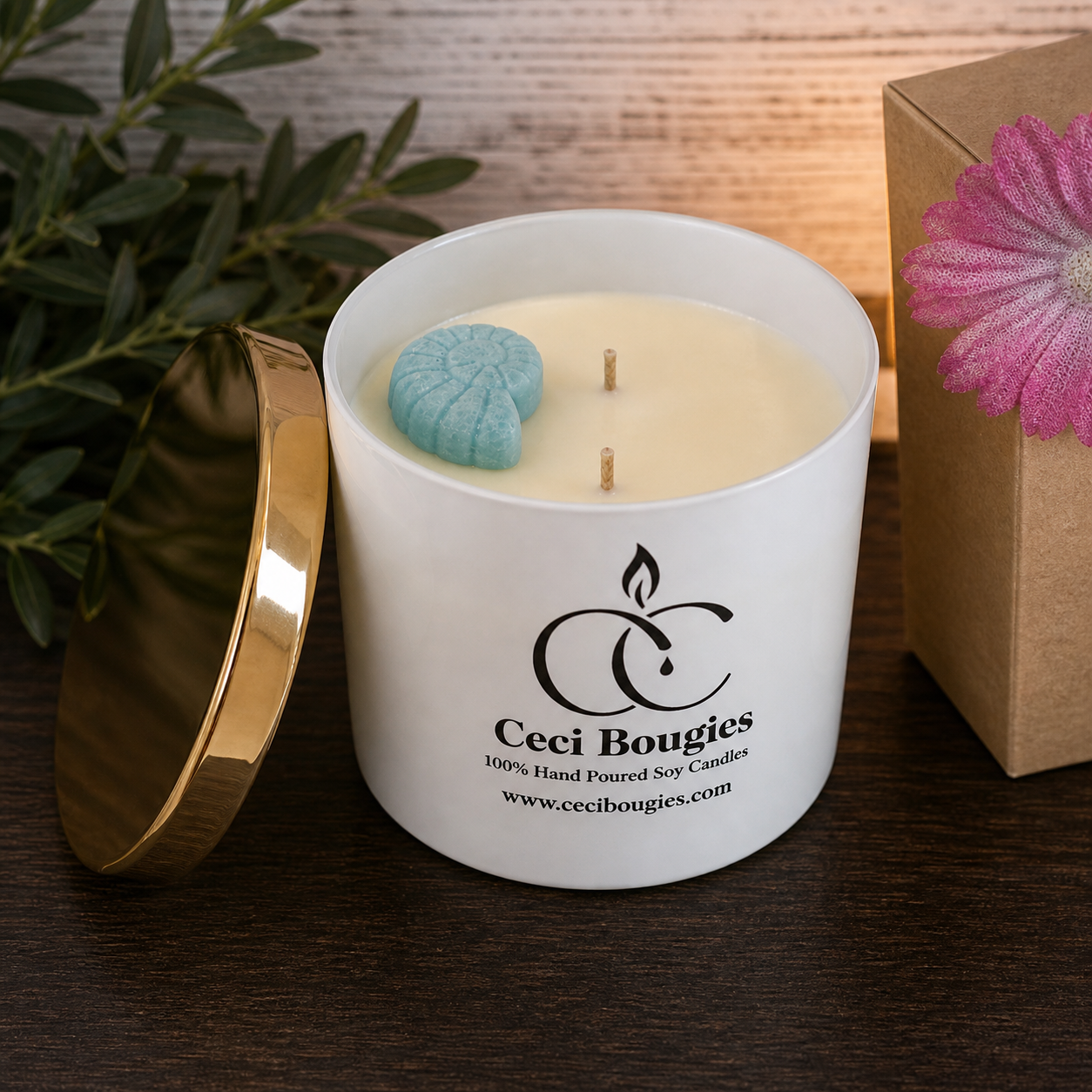 A white scented candle in a jar labeled "Ceci Bougies" with a gold lid leaning against it, pink flower decoration on the side, placed on a wooden surface.