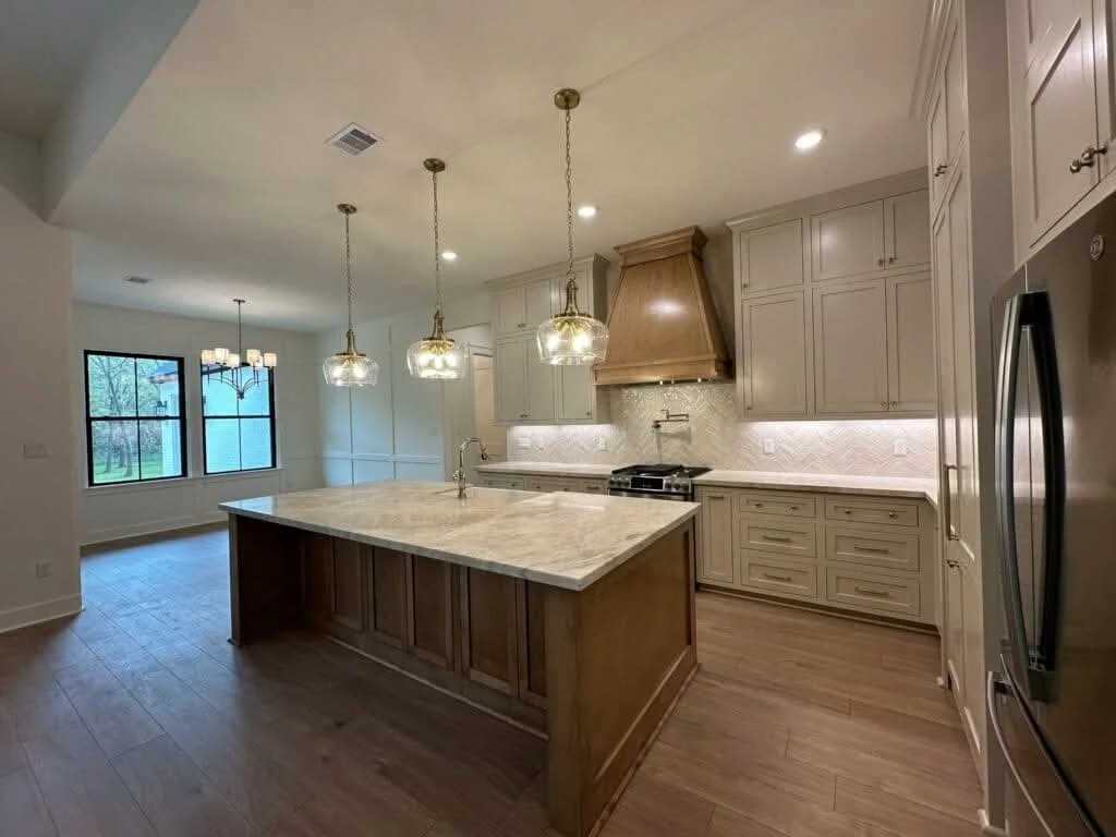 Modern kitchen with a large island, pendant lights, white cabinets, a stainless steel refrigerator, and a dining area with a chandelier and large windows.