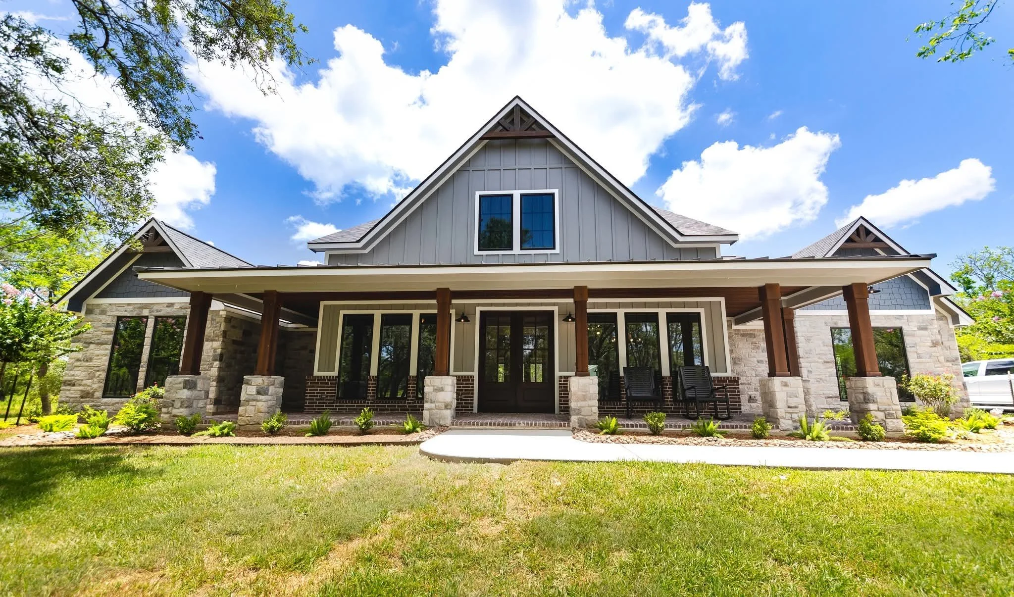 A modern house with a stone and siding exterior, a large porch with wooden columns, and a well-maintained lawn under a blue sky with clouds.