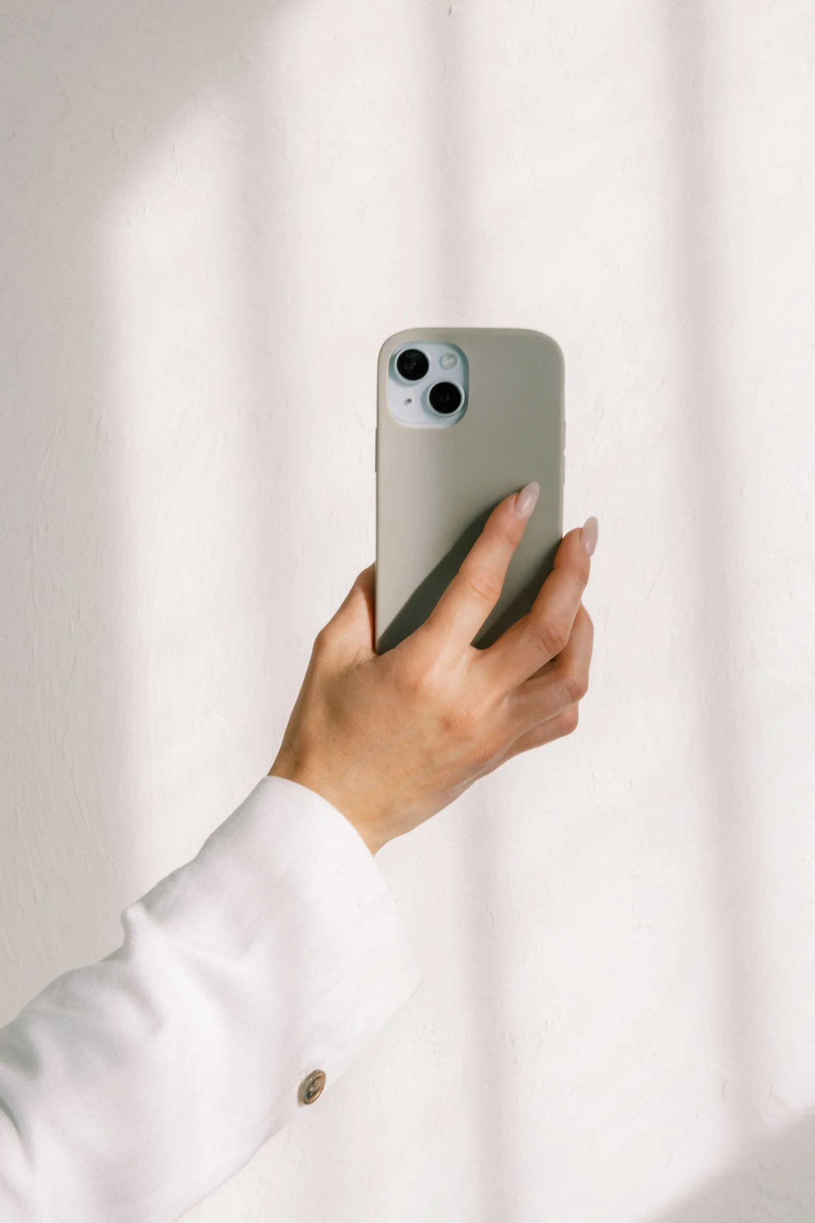 Person holding a smartphone with a white case, taking a selfie in front of a light-colored curtain.