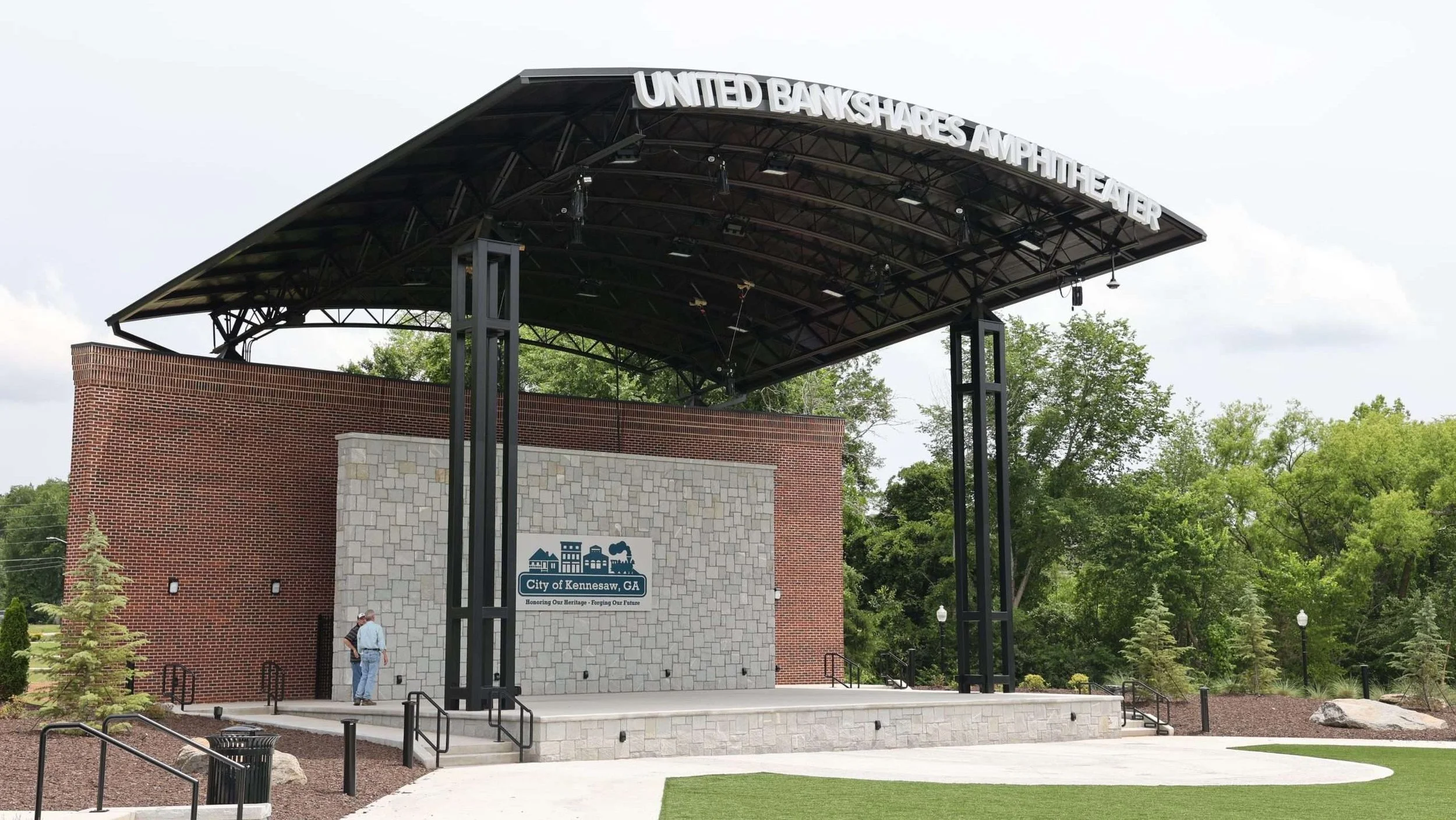 Entrance to the United Bankshares Amphitheater with a modern metal and wood canopy. The amphitheater is in the City of Kennesaw, Georgia, with a brick and stone facade, greenery, and trees in the background.