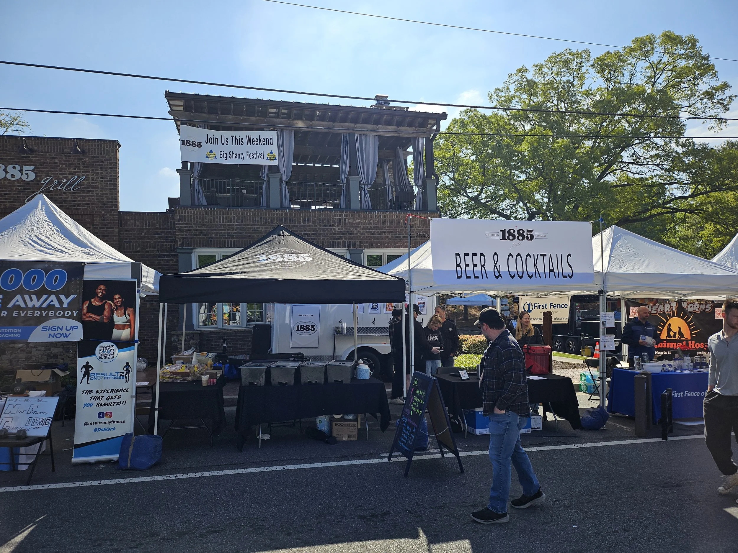 Street scene at a festival with white tents, one labeled 'BEER & COCKTAILS,' and people walking by. A sign promotes the Big Shanty Festival, and a second level above has a banner inviting people to join for the weekend.