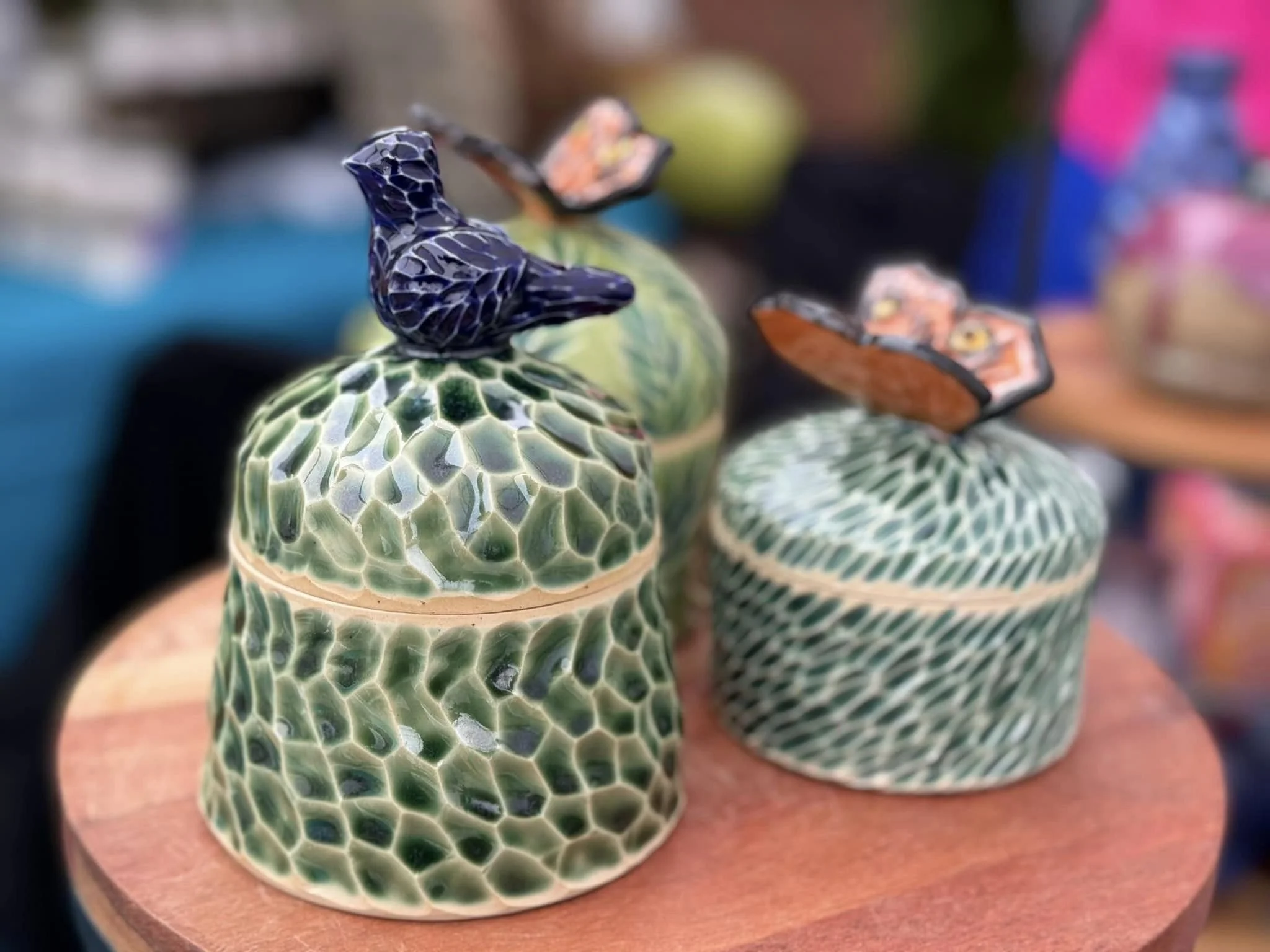 Two decorative ceramic boxes with lids featuring bird and butterfly figurines on top, placed on a round wooden surface.