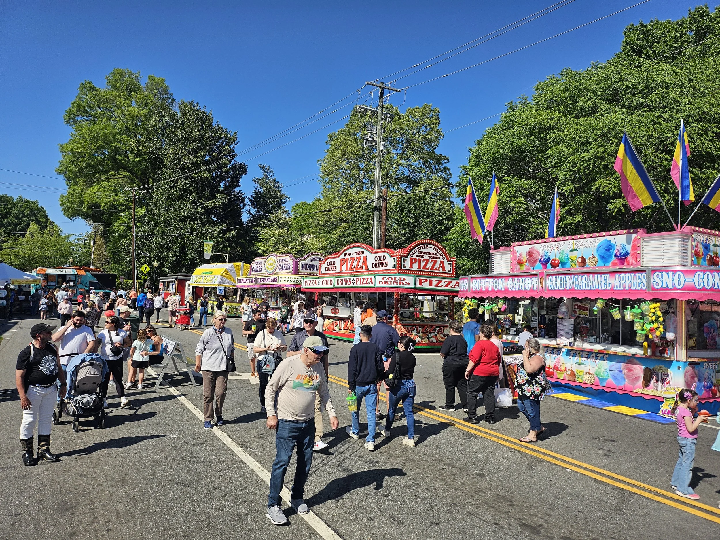 People walking along a street fair with food stalls, including pizza, cotton candy, and carnival treats. Colorful flags are displayed, and large trees are visible in the background under a bright blue sky.