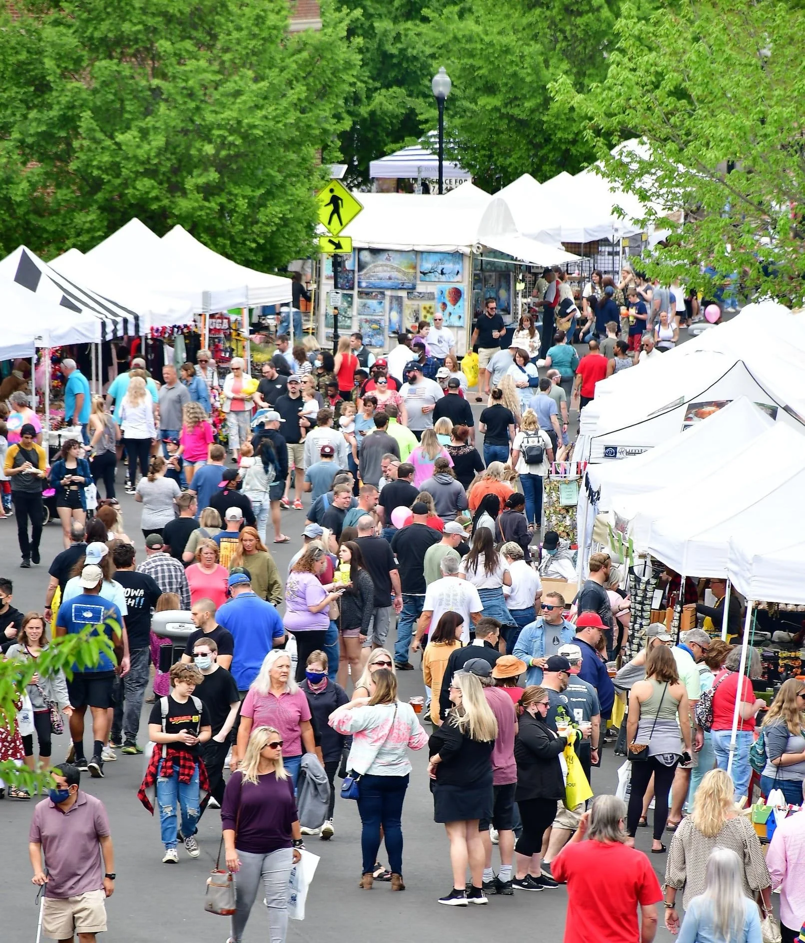Crowd of people walking and shopping at an outdoor market with white tents and trees in the background.