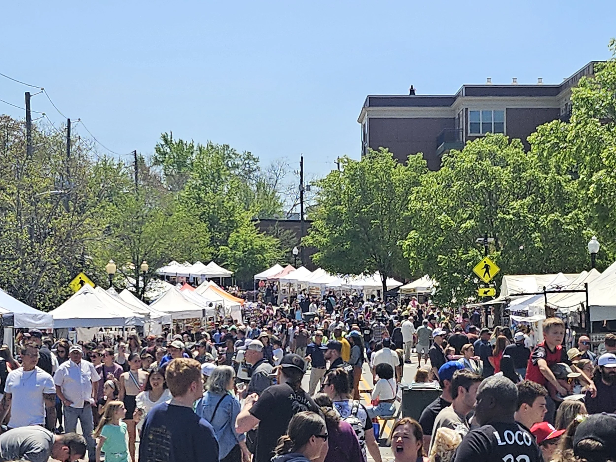 Crowd of people at an outdoor market with white tents, green trees, a blue sky, and buildings in the background.