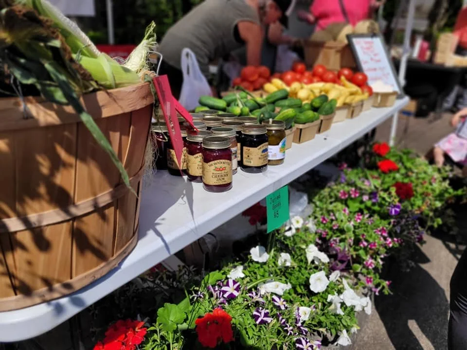 Table at farmers market with jars of jam, fresh vegetables like tomatoes and zucchinis, and colorful flowers below.