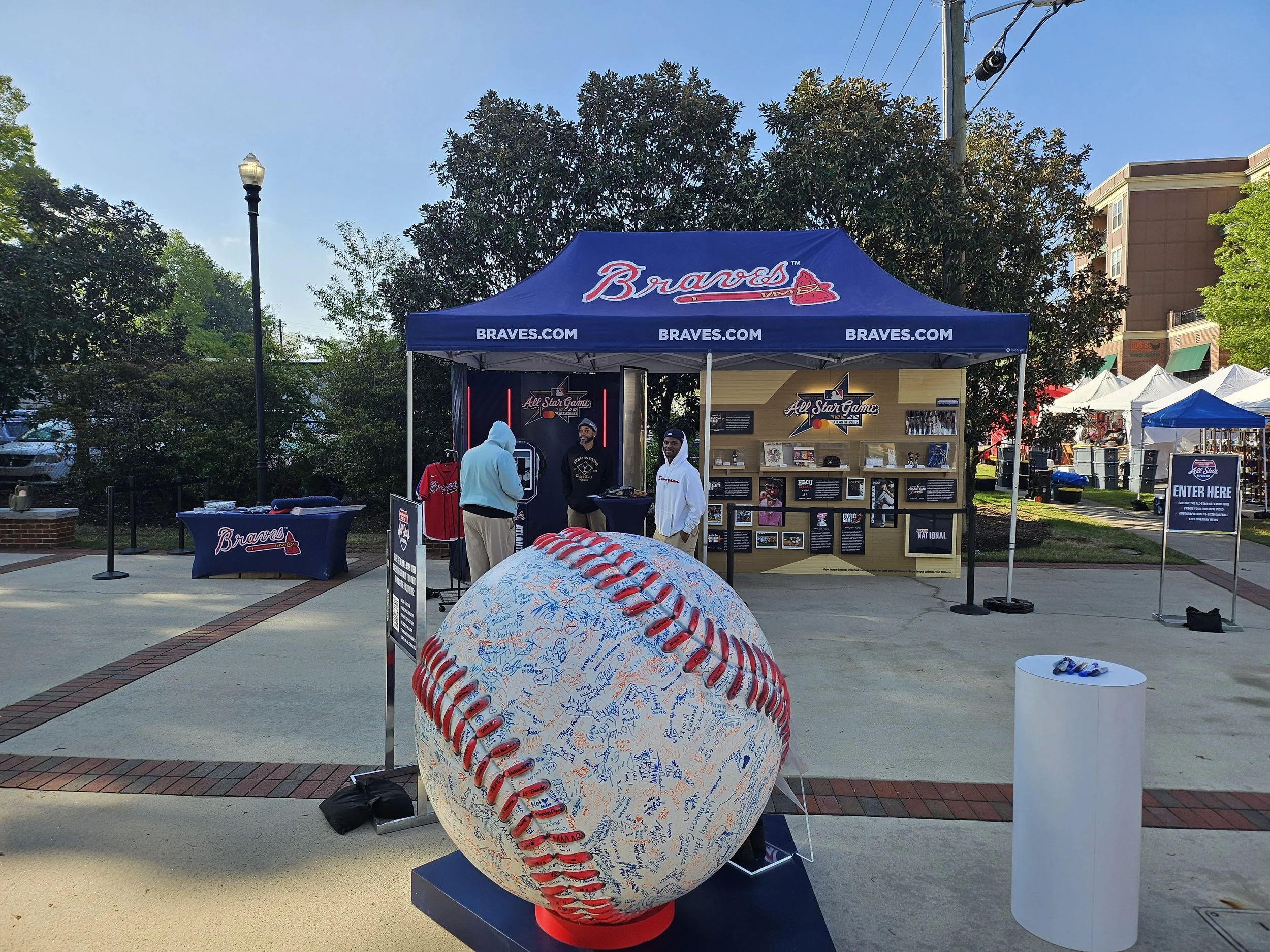 A large white baseball with red stitching, covered in signatures, displayed outside a Braves baseball team booth at an event. The booth has a blue canopy with 'Braves' written on it and multiple displays and merchandise related to the Atlanta Braves.
