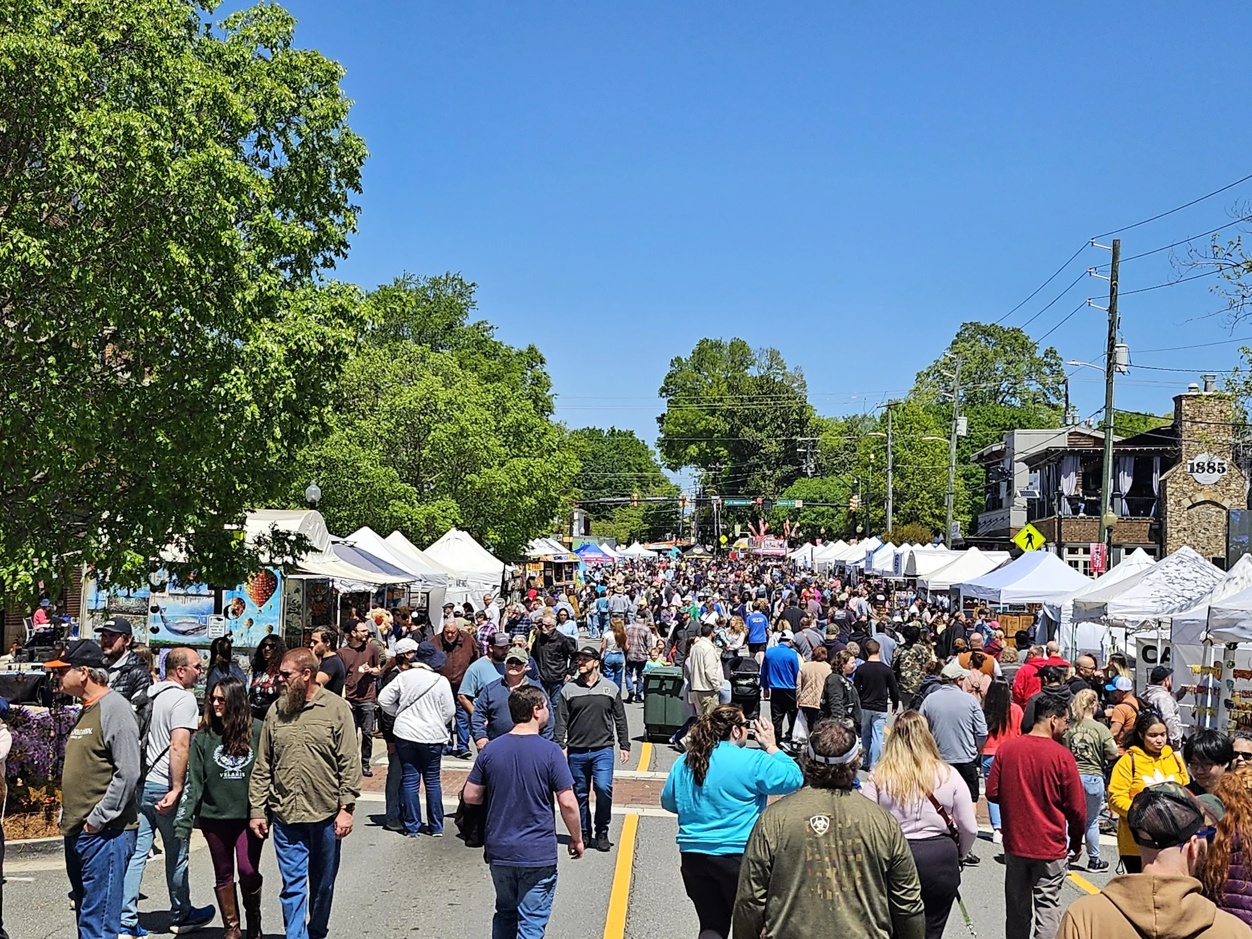 Crowd of people walking down a street lined with white tents and booths, with trees and buildings in the background under a clear blue sky.