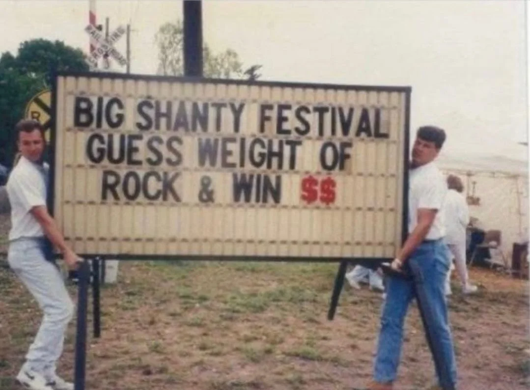Two men holding a large sign at the Big Shanty Festival, which encourages guessing the weight of rock and winning $5.