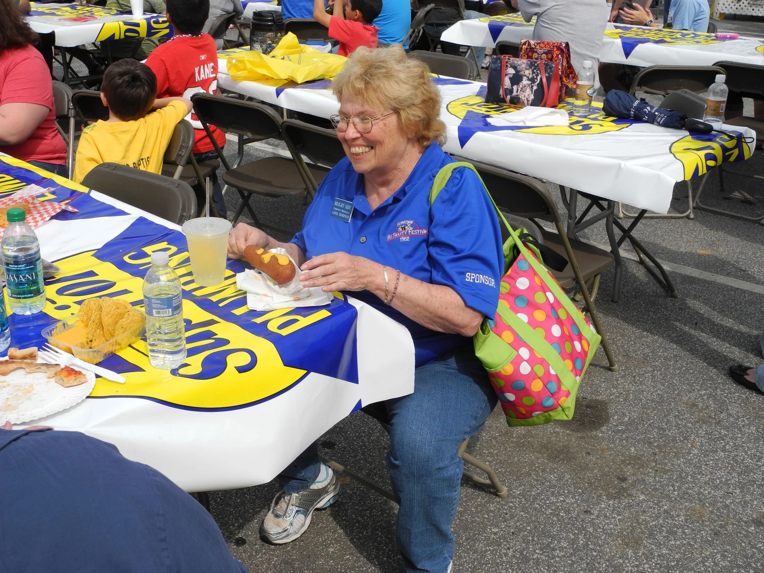 An older woman sitting at a table during a festival, smiling and holding a hot dog.