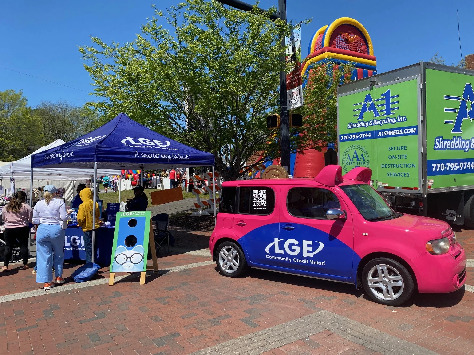 A community event featuring a pink and blue LGE Community Credit Union vehicle parked on a brick sidewalk next to a blue LGE tent booth with people gathered around. There is a green shredding truck nearby and an inflatable fun activity in the background with a large colorful arcade game structure.