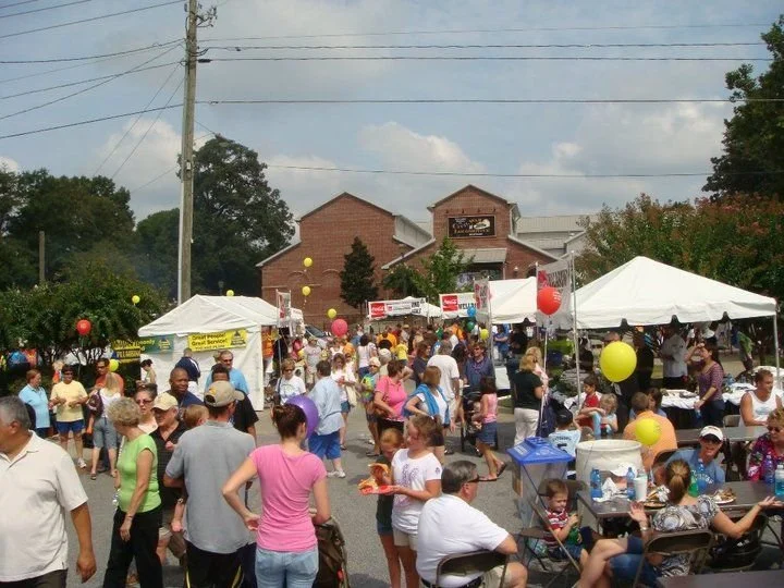 Crowd of people at outdoor festival with tents, tables, and balloons in a parking lot, with a brick building in the background.